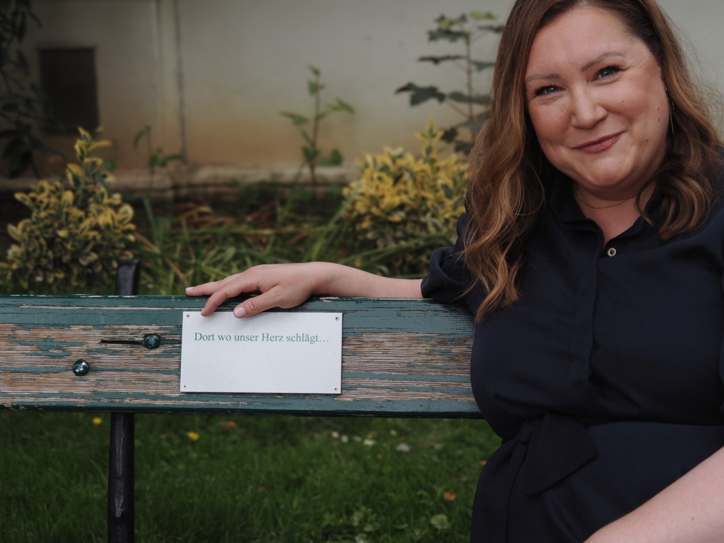 Eine Frau mit braunen, lockigen Haaren, die ein schwarzes Hemd trägt, lehnt an einer grau-grünen Parkbank, auf deren Rückenlehne ein Schild mit der Aufschrift "Dort, wo unser Herz schlägt..." hängt. Im Hintergrund sind Blumen und eine graue Mauer zu sehen.