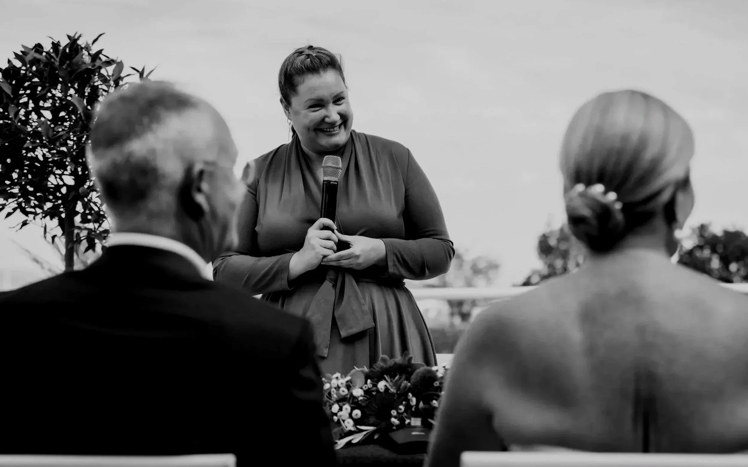 Eine lachende Frau hält ein Mikrofon und spricht vor einer Gruppe. Im Hintergrund sind Bäume, im Vordergrund sind mehrere Menschen zu sehen, die sich die Frau anhören.
