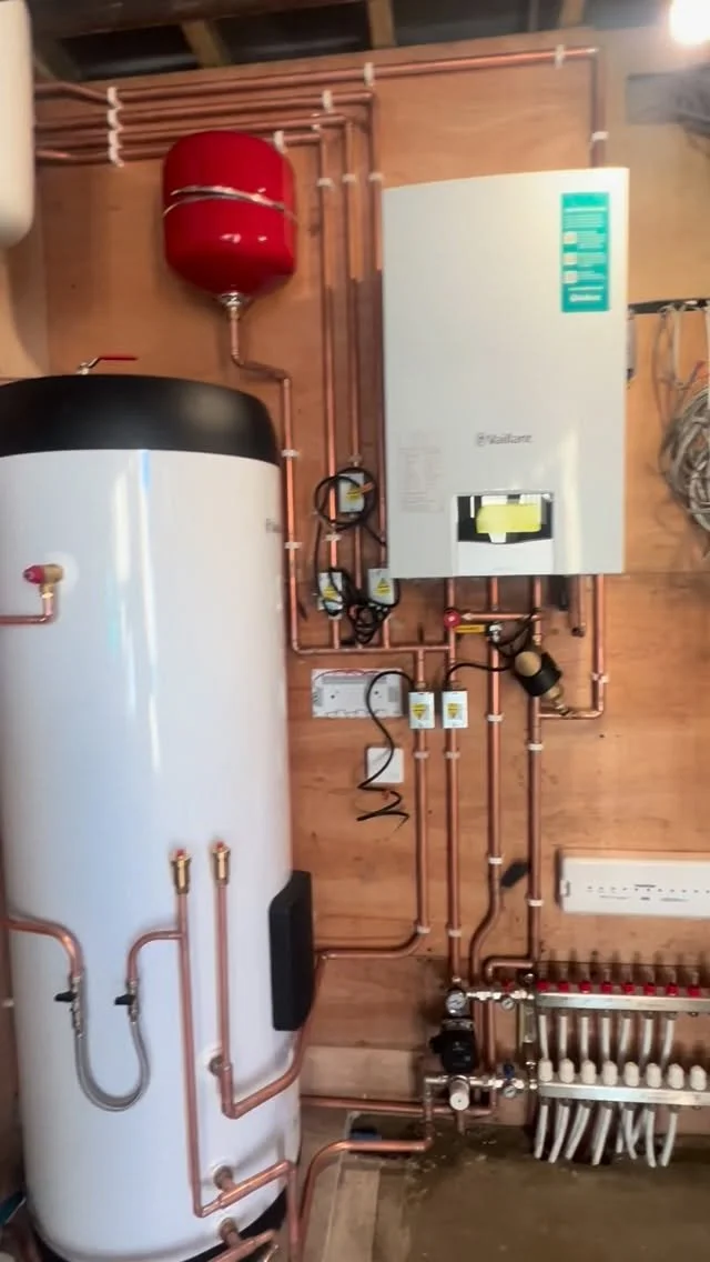 A mechanical room with a large white water heater, a red expansion tank above it, and a white boiler on the wall, all connected by copper pipes.