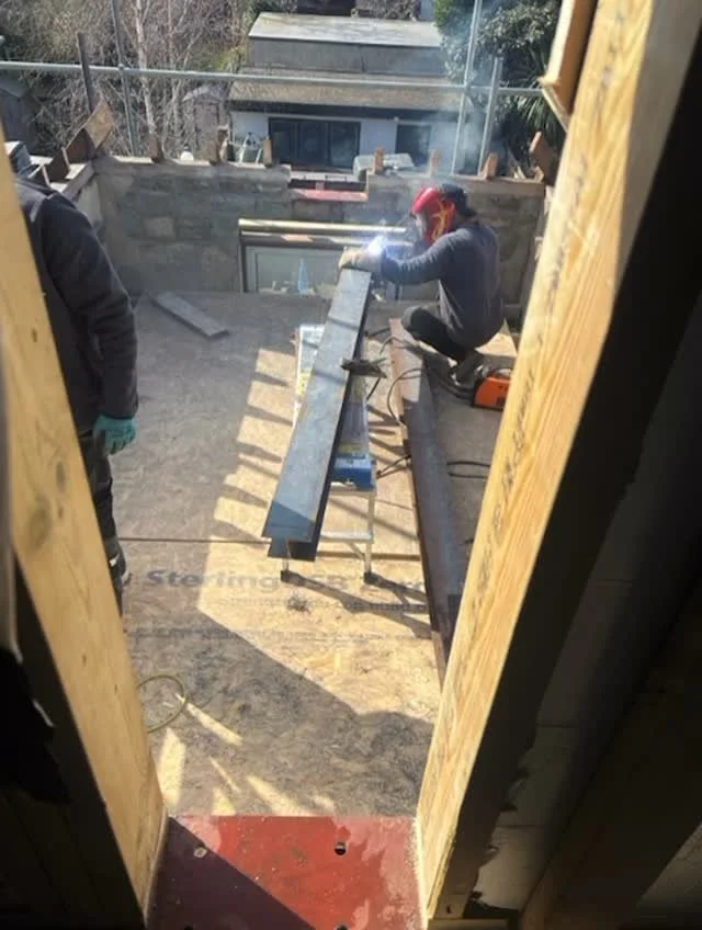 A construction worker wearing a red helmet and safety gloves is crouched on a wooden beam, using a welding machine to work on a metal pipe, on an outdoor roof or terrace. There are two workers, with the second worker partially visible on the left side, wearing gloves and dark clothing. The area is surrounded by a glass railing and has a concrete surface with some tools and a sawhorse.