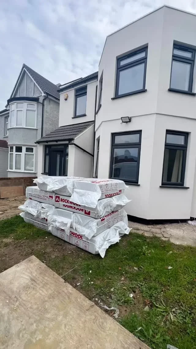 Stacks of insulation material in white plastic bags outside a modern white house with black window frames, on a patch of grass and dirt.