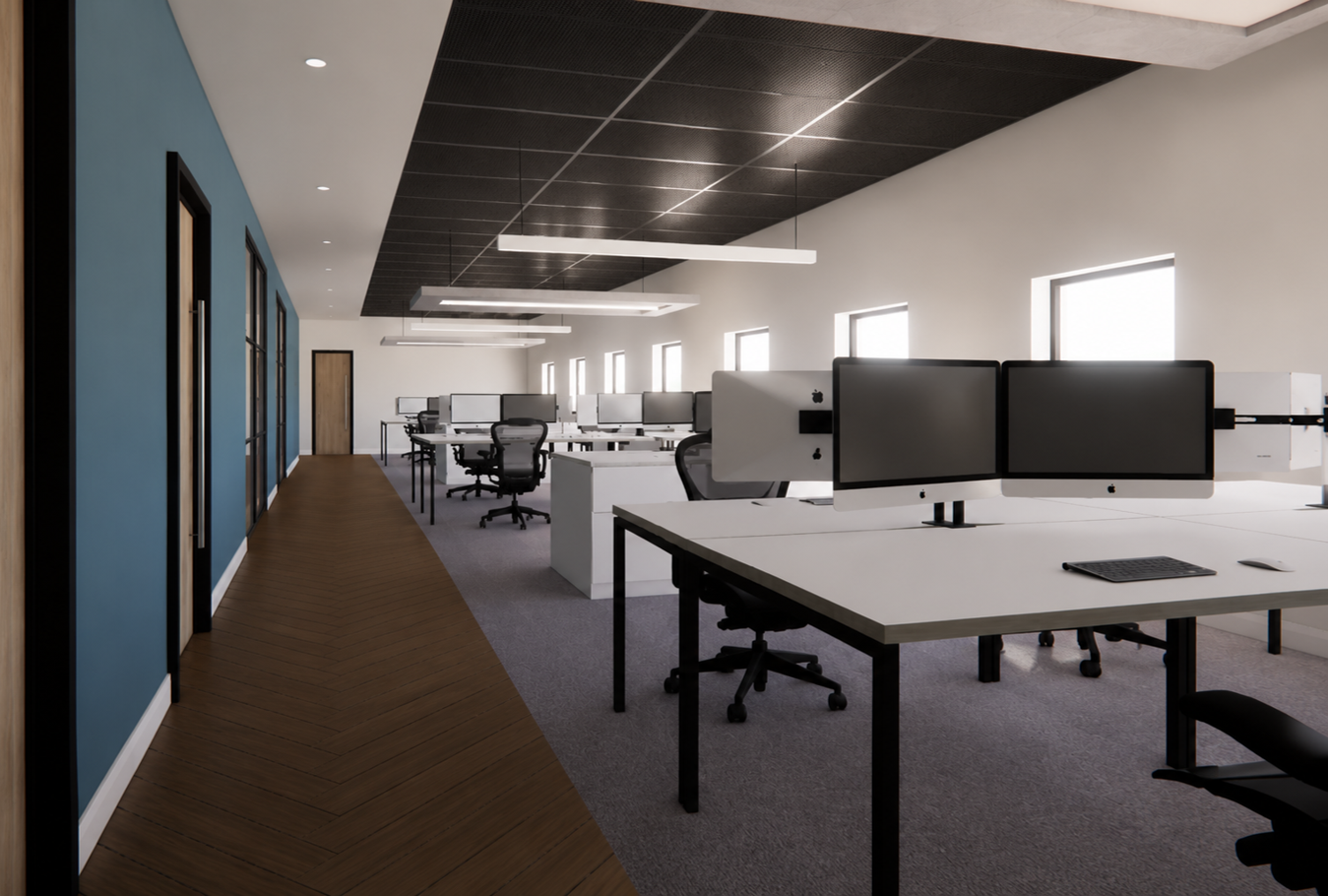 Modern office workspace with multiple desks, dual computer monitors, ergonomic chairs, and large windows letting in natural light, featuring a black ceiling with suspended lights.