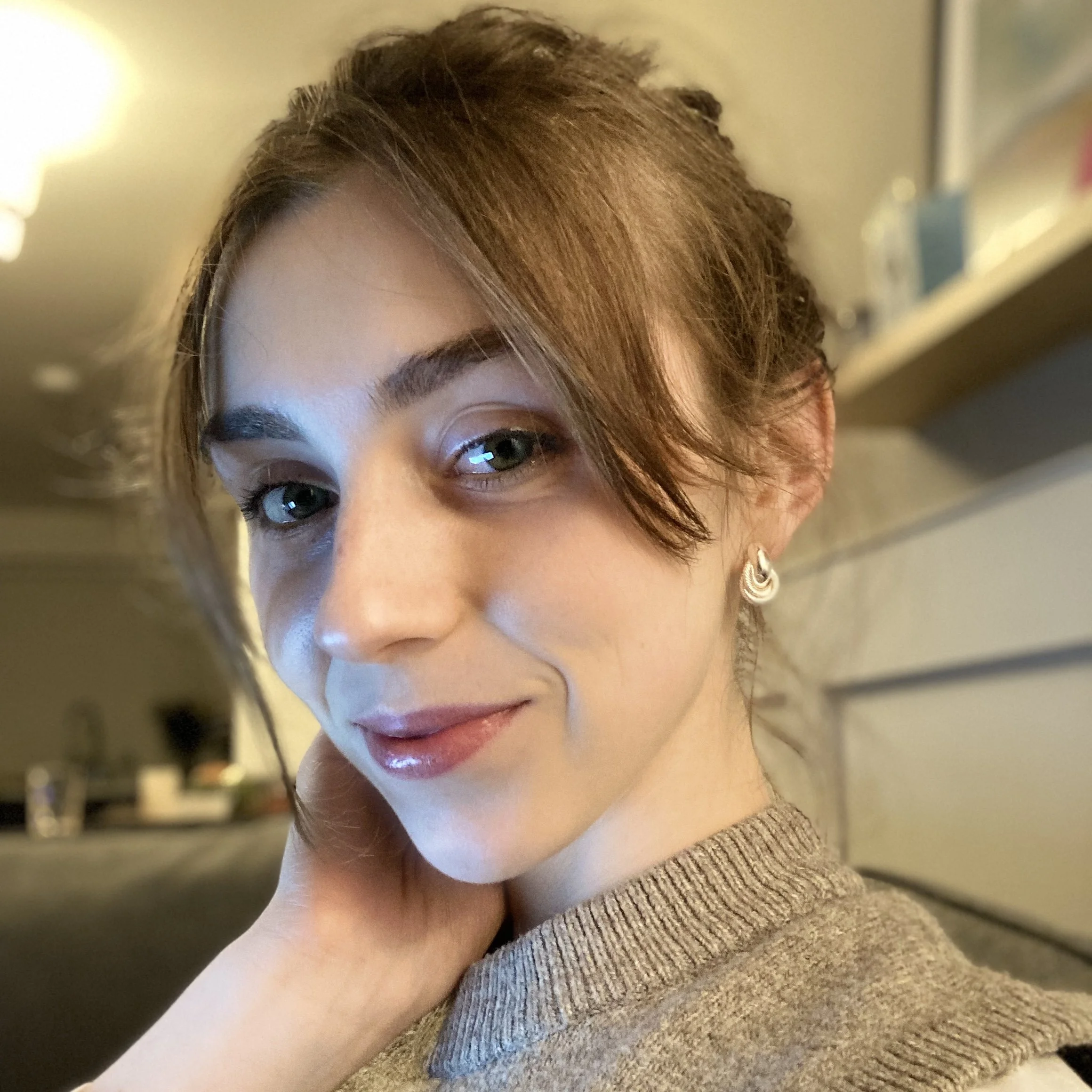 Close-up of a young woman's face with brown hair and green eyes, wearing an earring, with a beige sweater and a blurred background.
