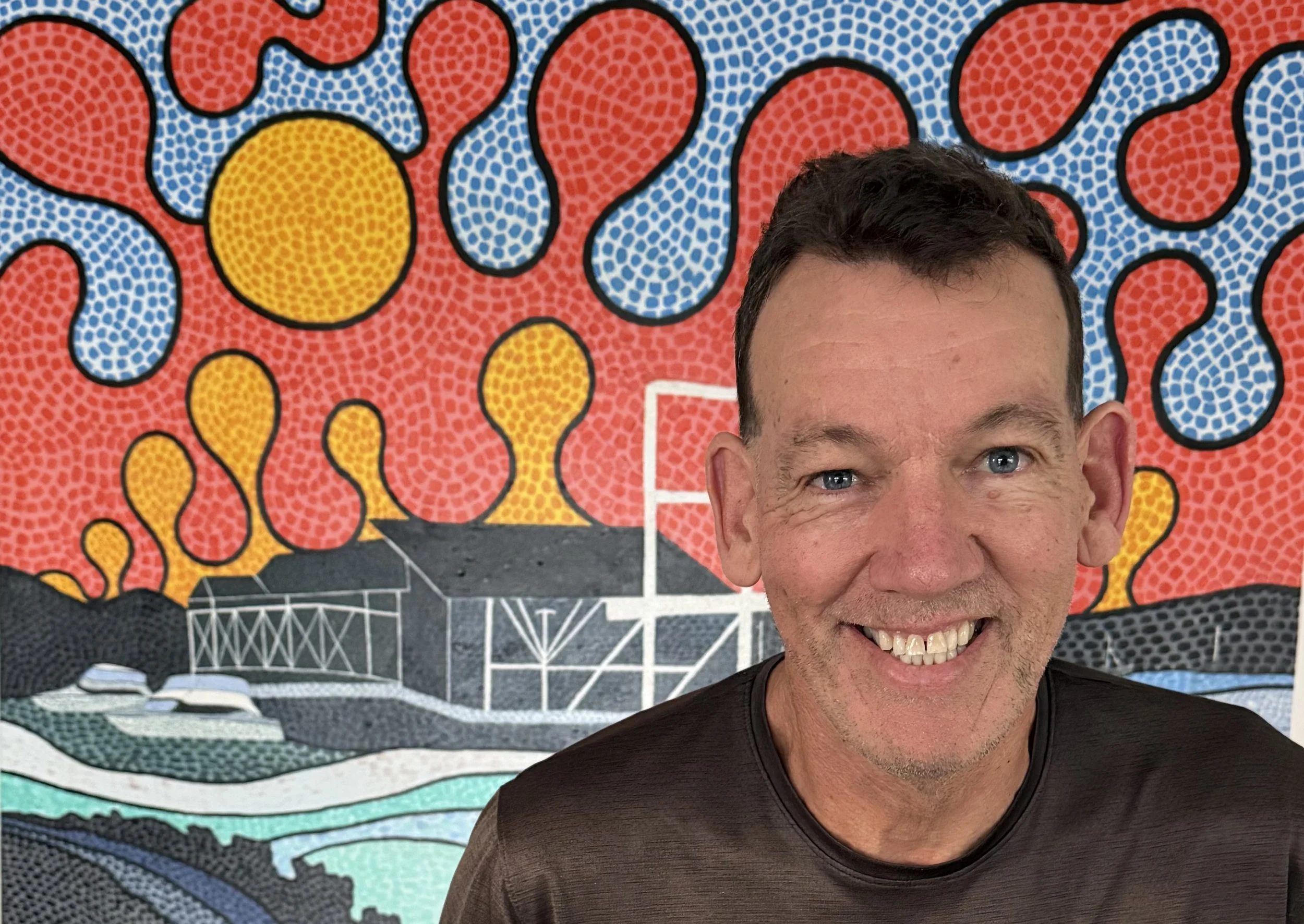 A smiling man with short dark hair and blue eyes standing in front of a colorful, abstract mural featuring bright red, yellow, blue, green, and black shapes.