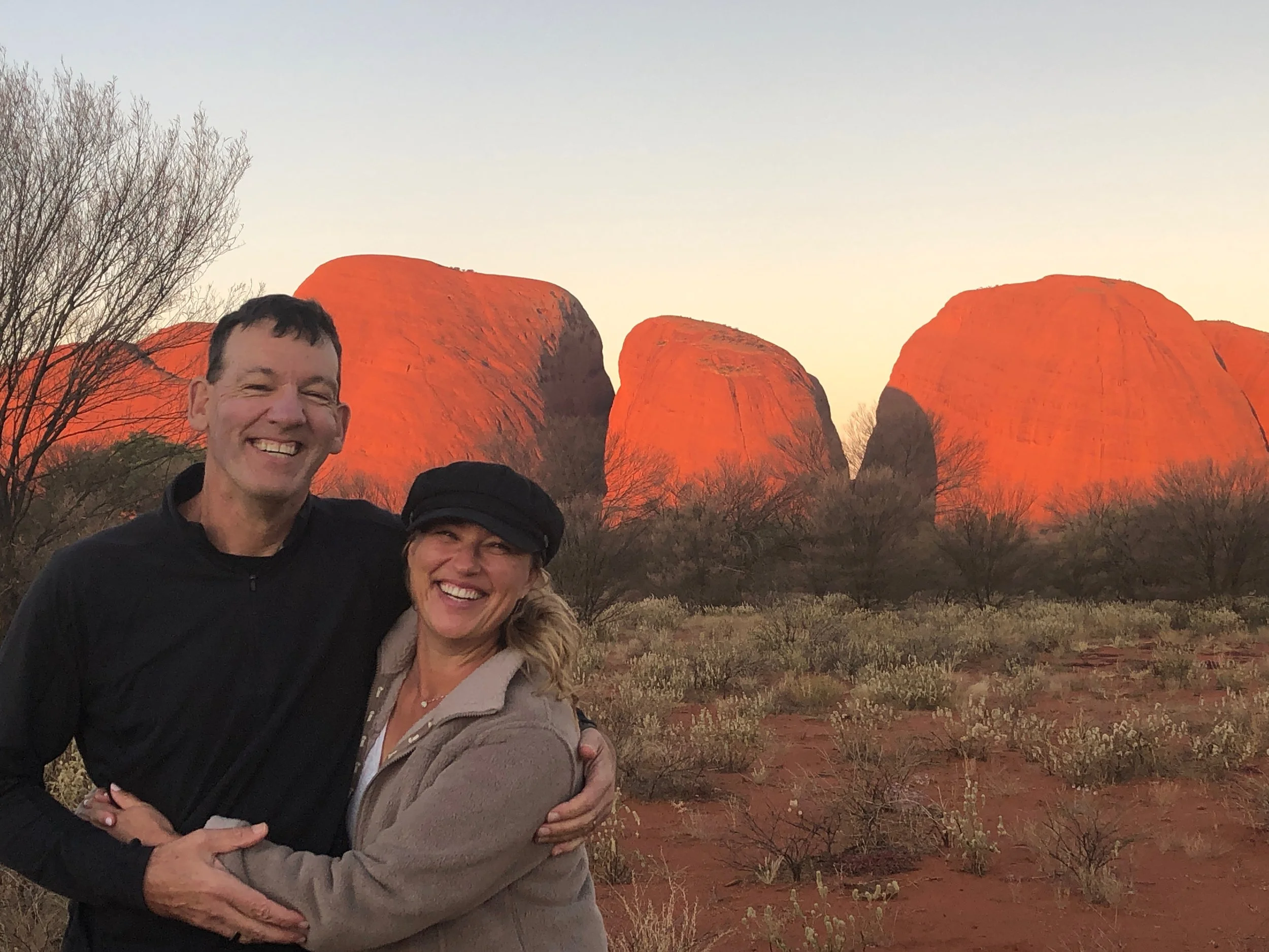 A group of people smiling and hugging with large red rock formations in the background during sunset or sunrise.