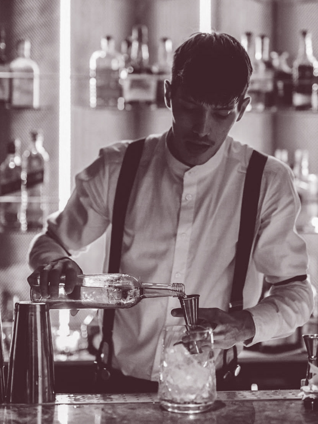 A bartender pouring a clear liquid into a glass at a bar, surrounded by bottles and barware, in black and white.