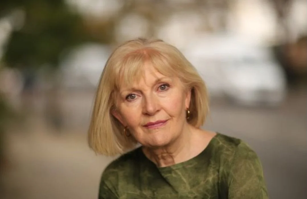 Close-up of an elderly woman with blonde hair, wearing a green patterned top, smiling softly outdoors.