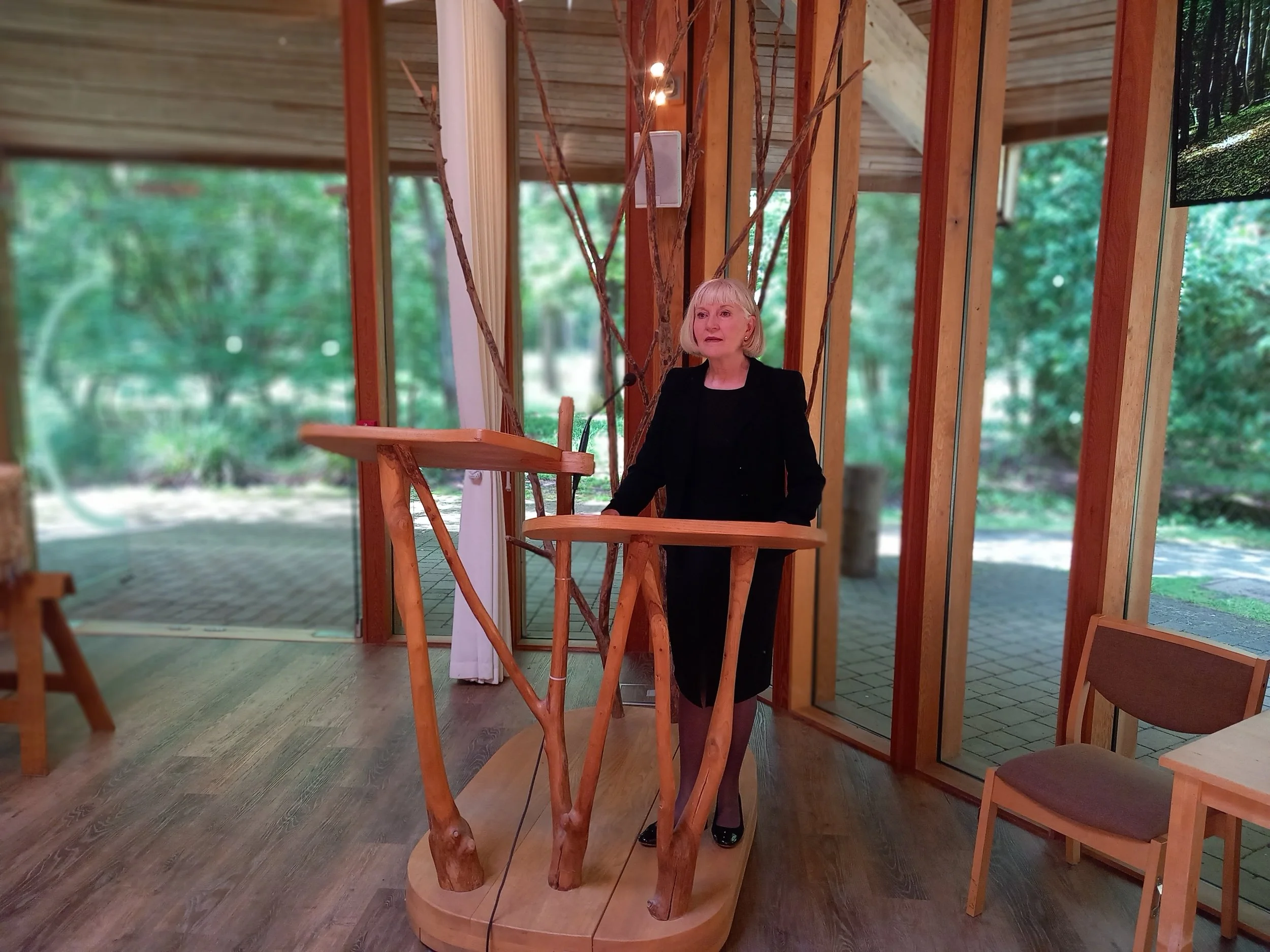 Woman with blonde hair wearing a black blazer and skirt, standing inside a wooden structure with trees visible outside.
