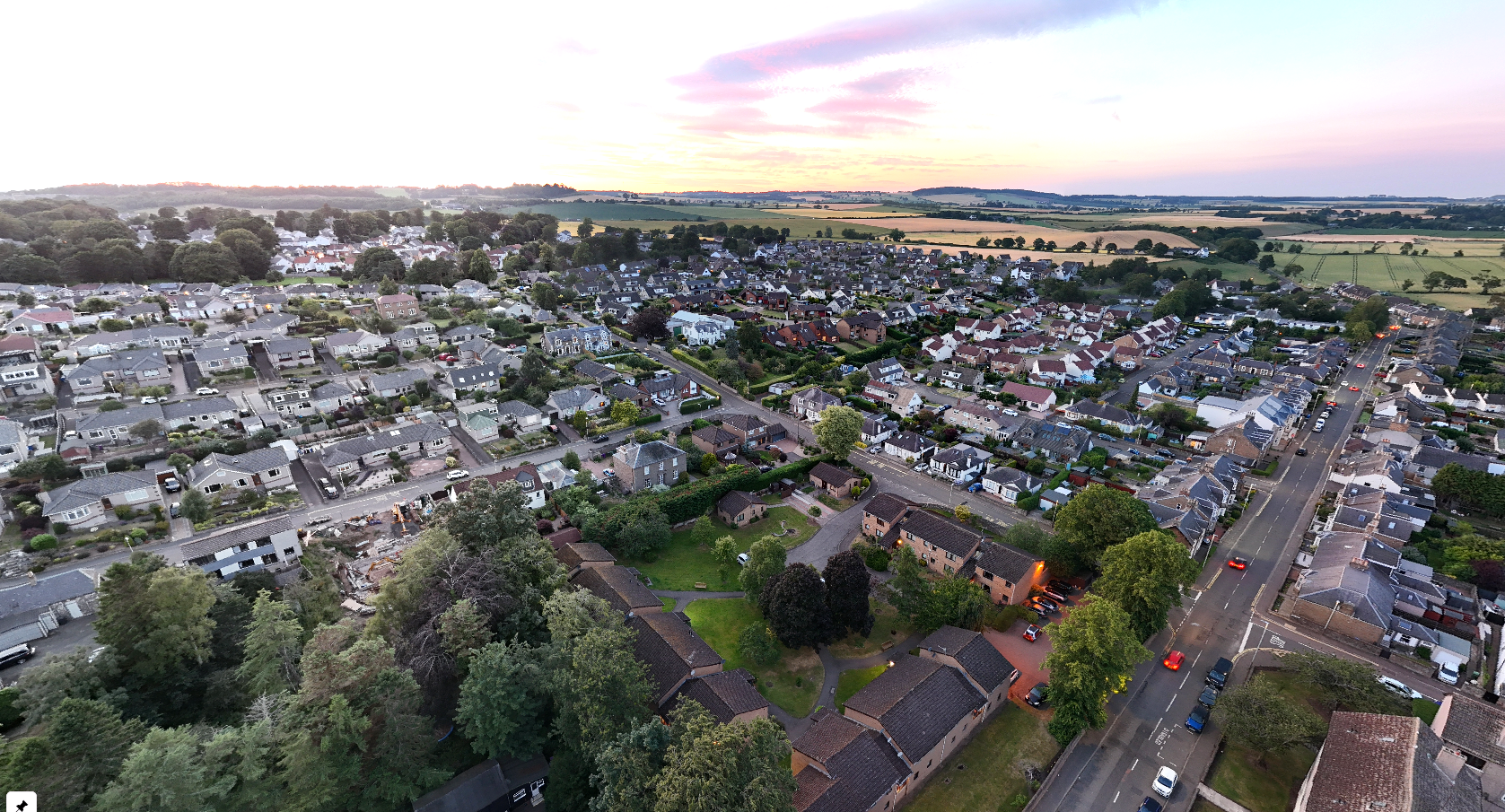 Aerial view of Monifieth at sunset with houses, roads, trees, and green fields in the background.