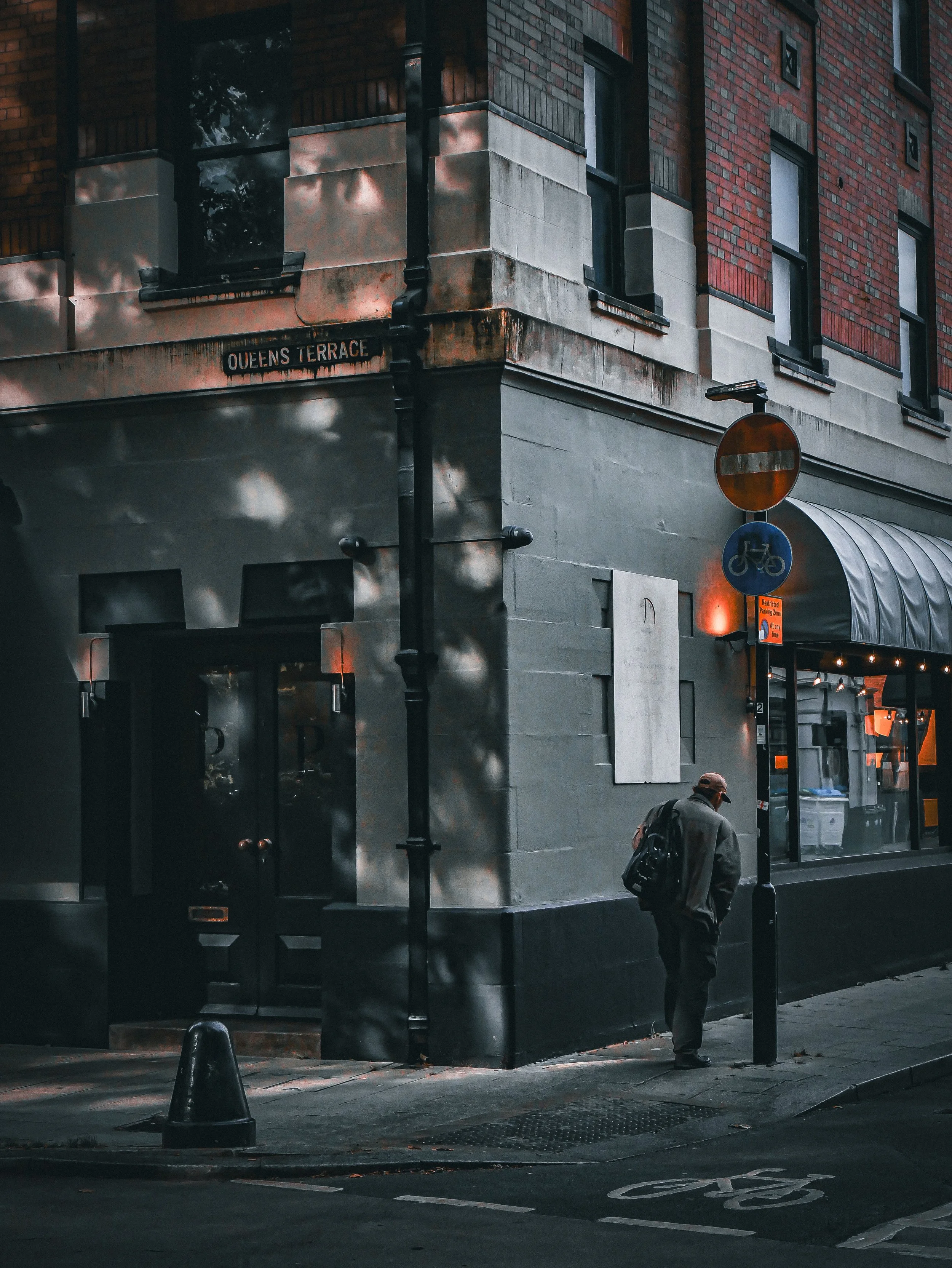 A man with a backpack walking on the sidewalk next to a building with a sign that reads "Queens Terrace." There are traffic cones, a no-entry sign, and a bike lane marking on the street. The scene appears to be in the evening or early morning with di