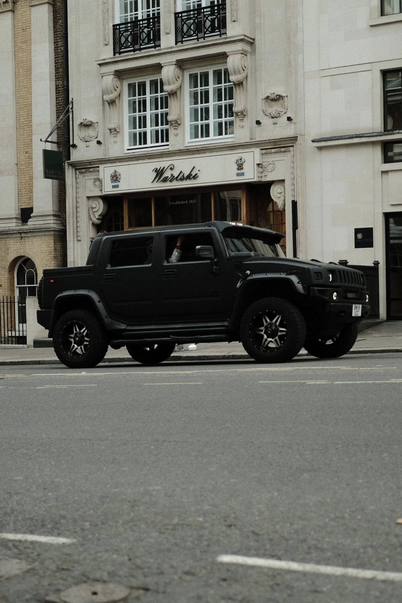 A black Humvee vehicle parked on a city street in front of a historic building with ornate architectural details, large windows, and a sign that reads "Wartski".