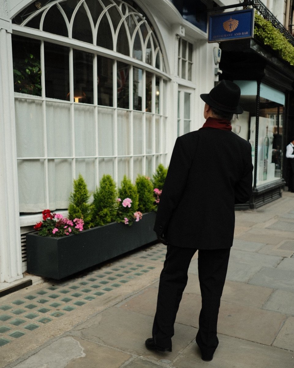 A person dressed in a black suit, black hat, and red scarf standing on a city sidewalk and looking at a restaurant storefront decorated with potted plants and flowers.