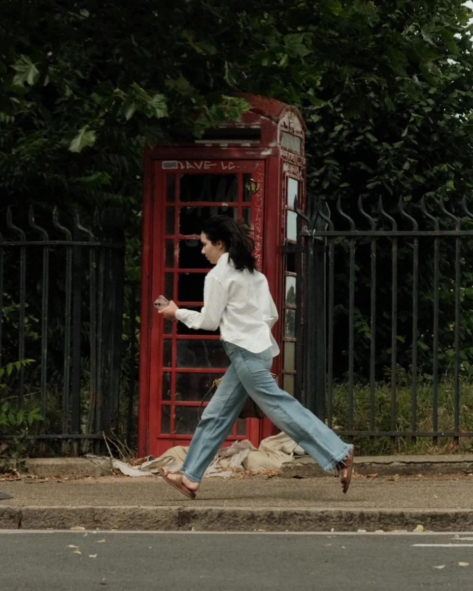 A woman walking quickly on a sidewalk while looking at her phone, with an old red British telephone booth and green foliage in the background.