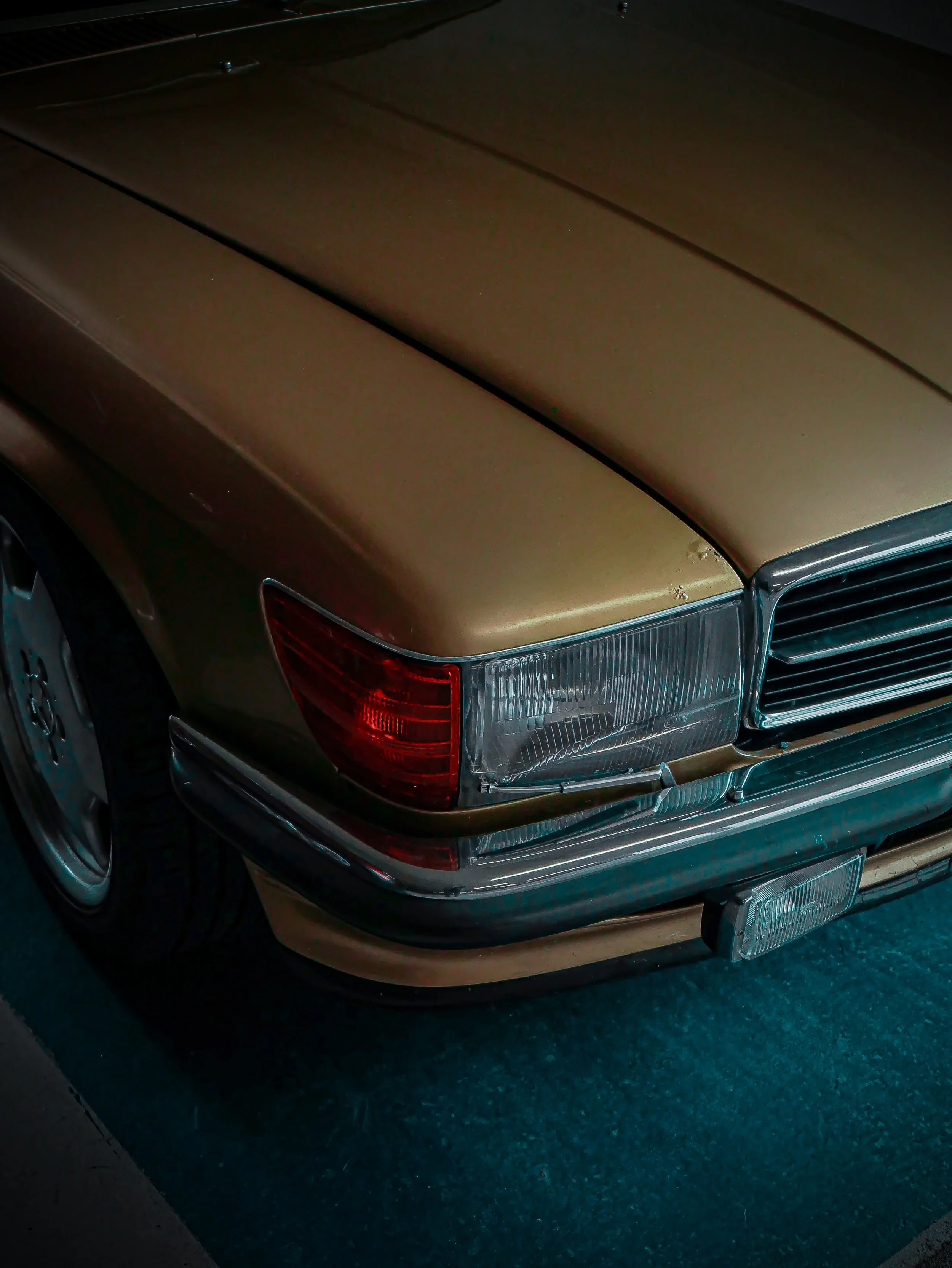 Close-up of the front corner of a vintage gold-colored car, showing part of the front grille, headlight, and bumper.