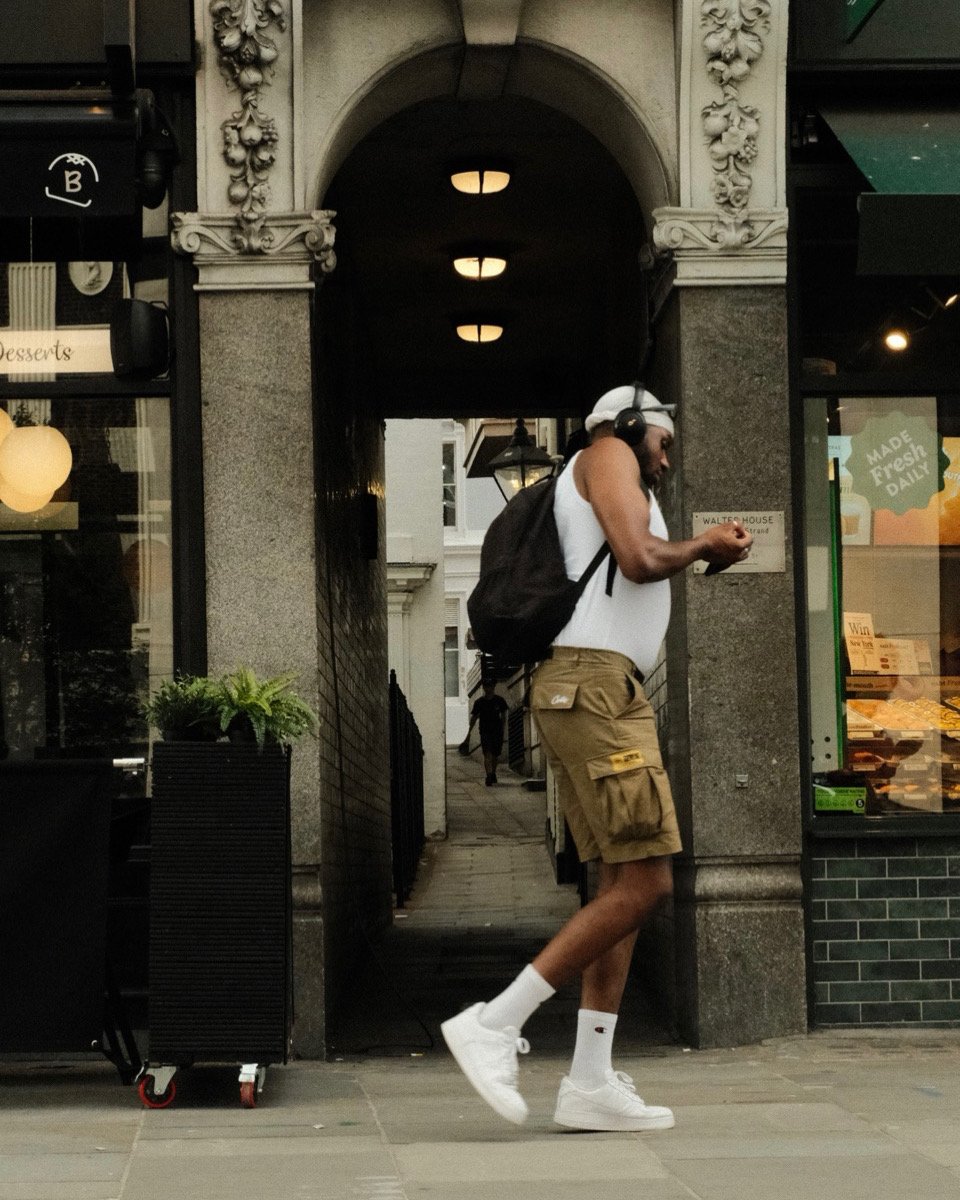 A man with a backpack, wearing headphones, a white tank top, khaki shorts, white socks, and white sneakers, walking past a building with an archway and using his phone.
