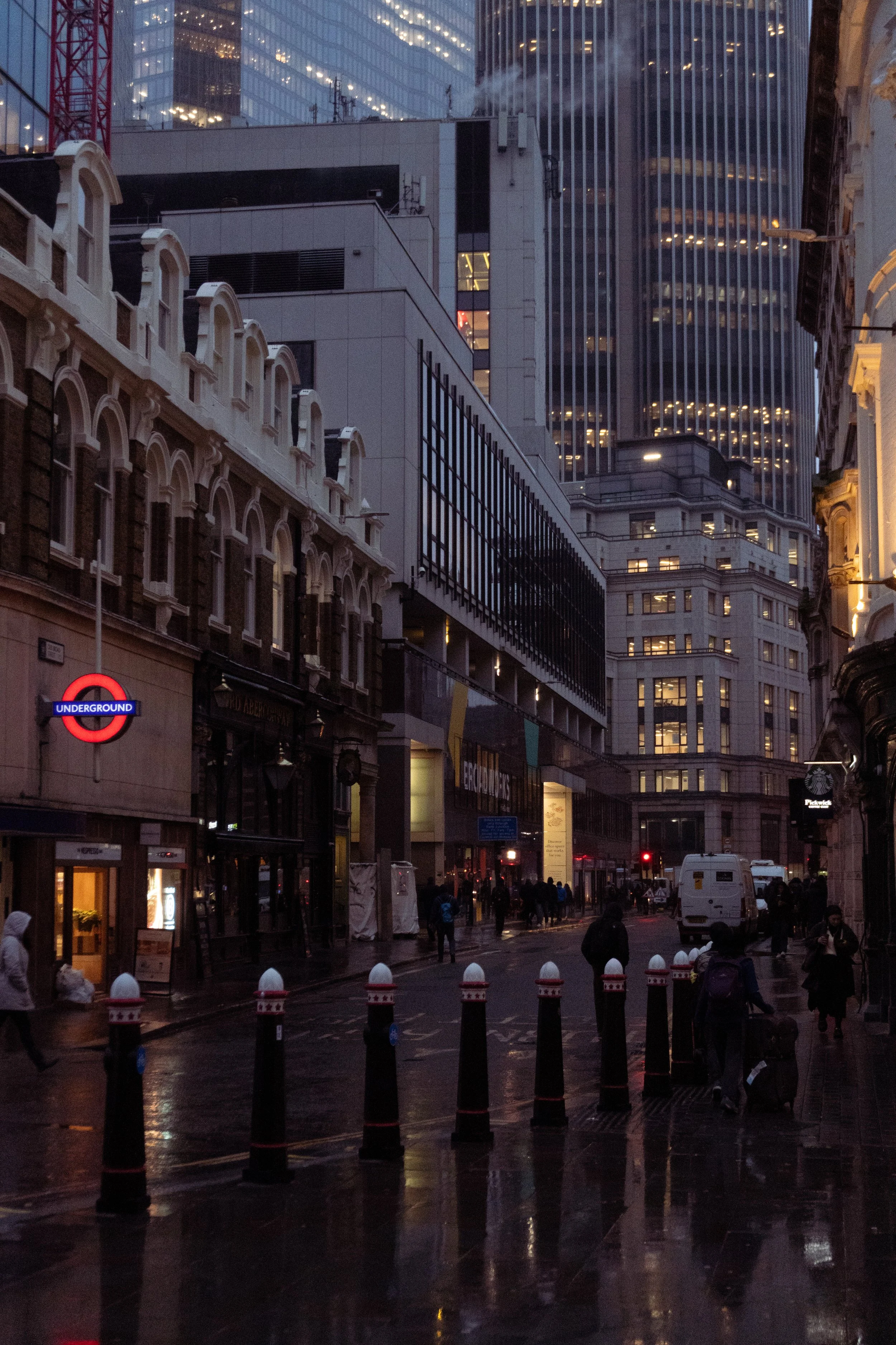 City street scene at dusk with high-rise buildings, shops, and pedestrians on wet pavement, with rain reflecting city lights.