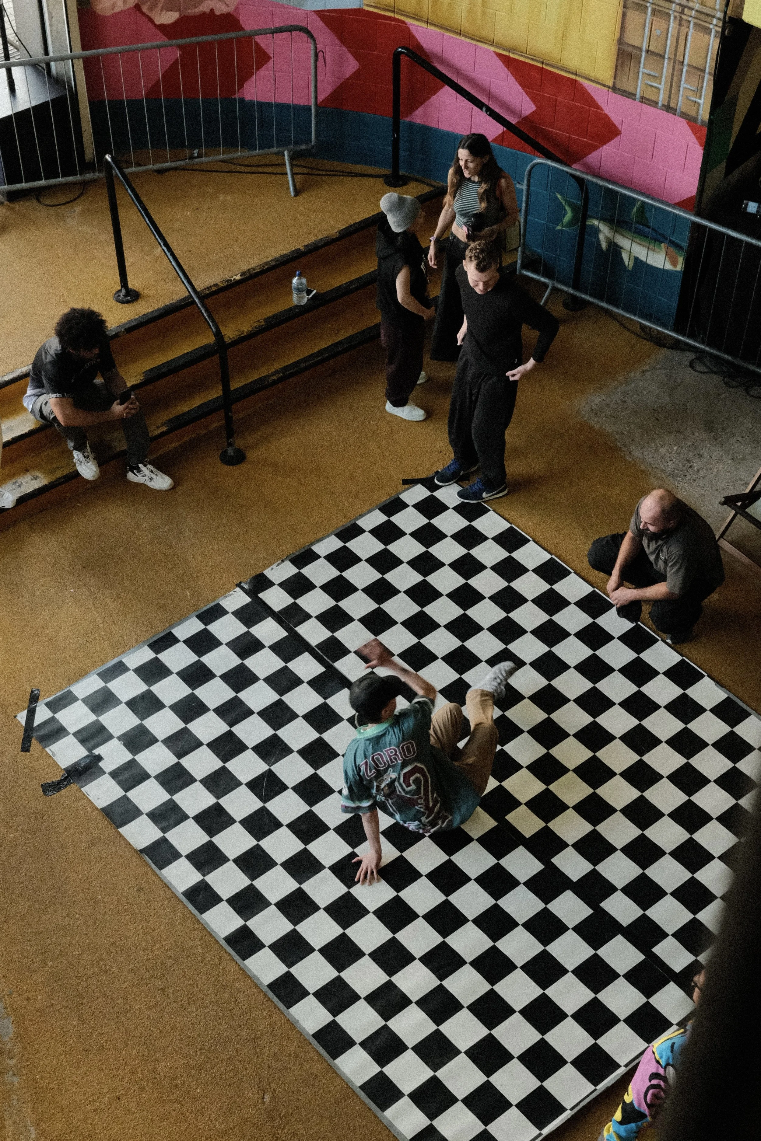 A group of people sitting and standing around a black-and-white checkered chessboard on the floor in a colorful room.
