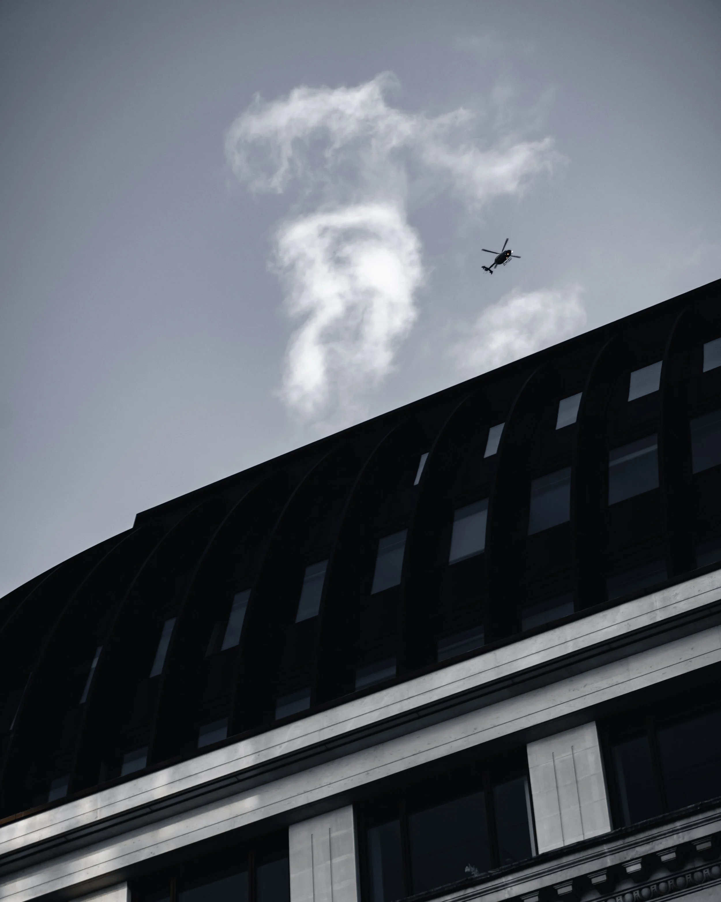 A helicopter flying near a modern multi-story building with dark and light-colored exterior, multiple windows, and a curved architectural design.