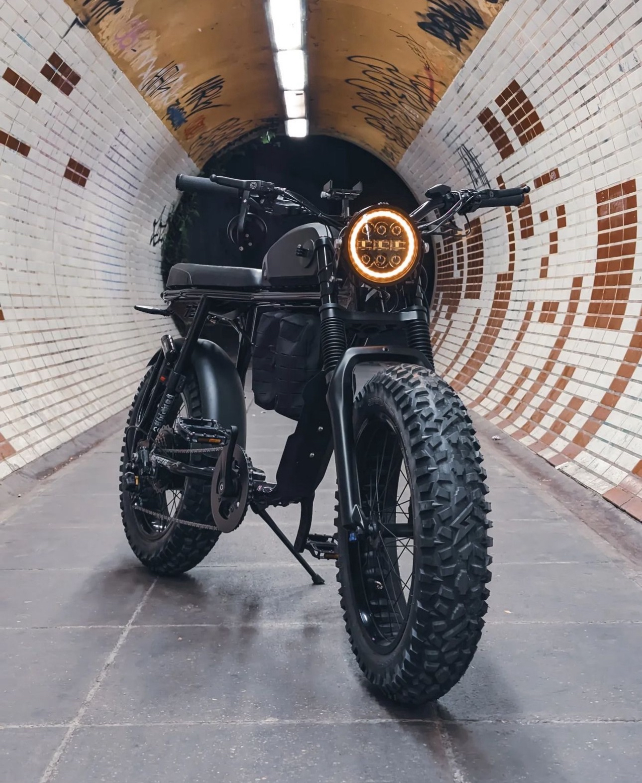 A black electric motorcycle with off-road tires parked inside a tiled underground tunnel with graffiti on the walls and ceiling, and a long fluorescent light fixture overhead.