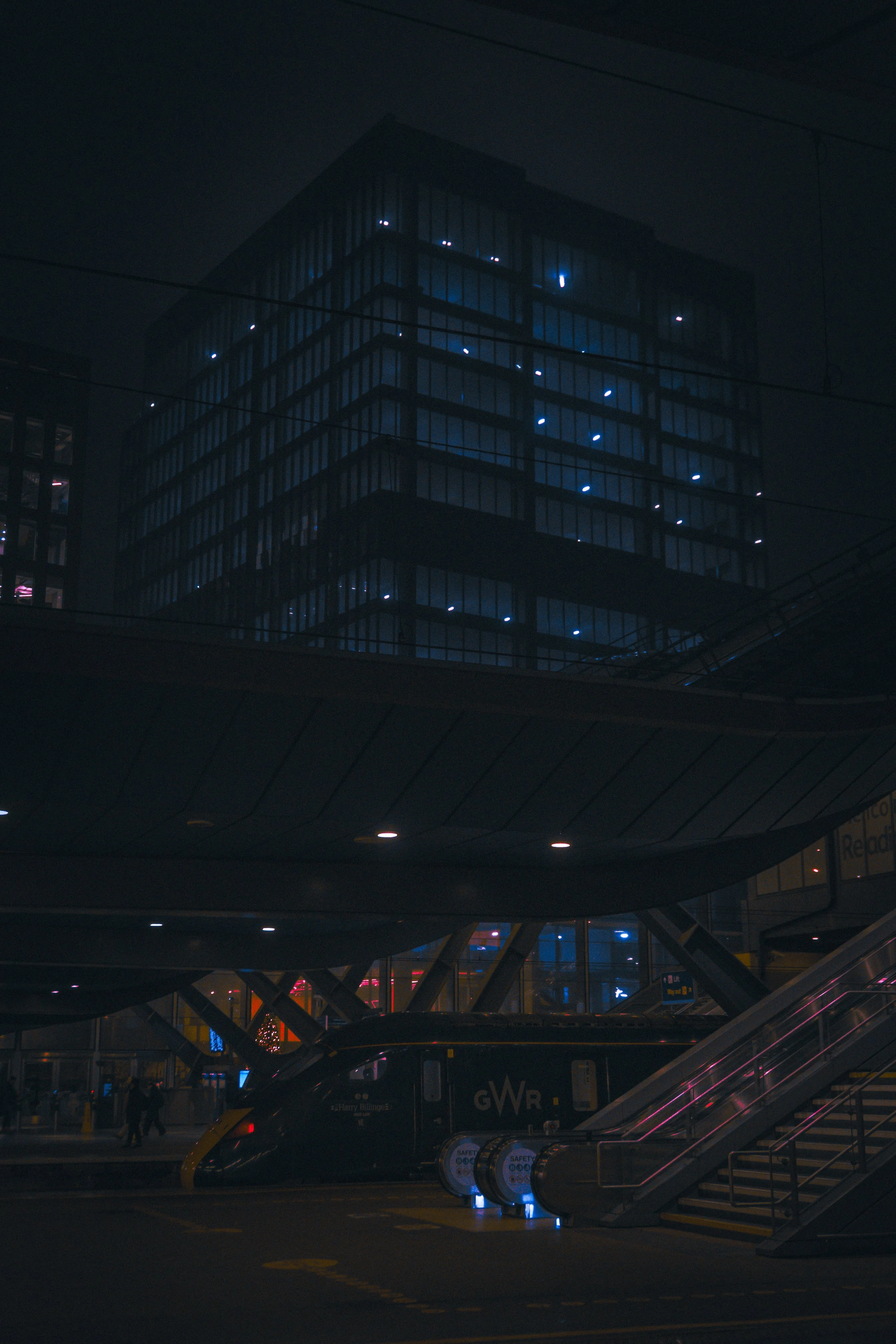 Nighttime scene at an airport with a dark building featuring blue lights inside. In the foreground, there are people walking, a bus with the GWR logo, an escalator, and safety information signs, with parking and airport terminal visible.