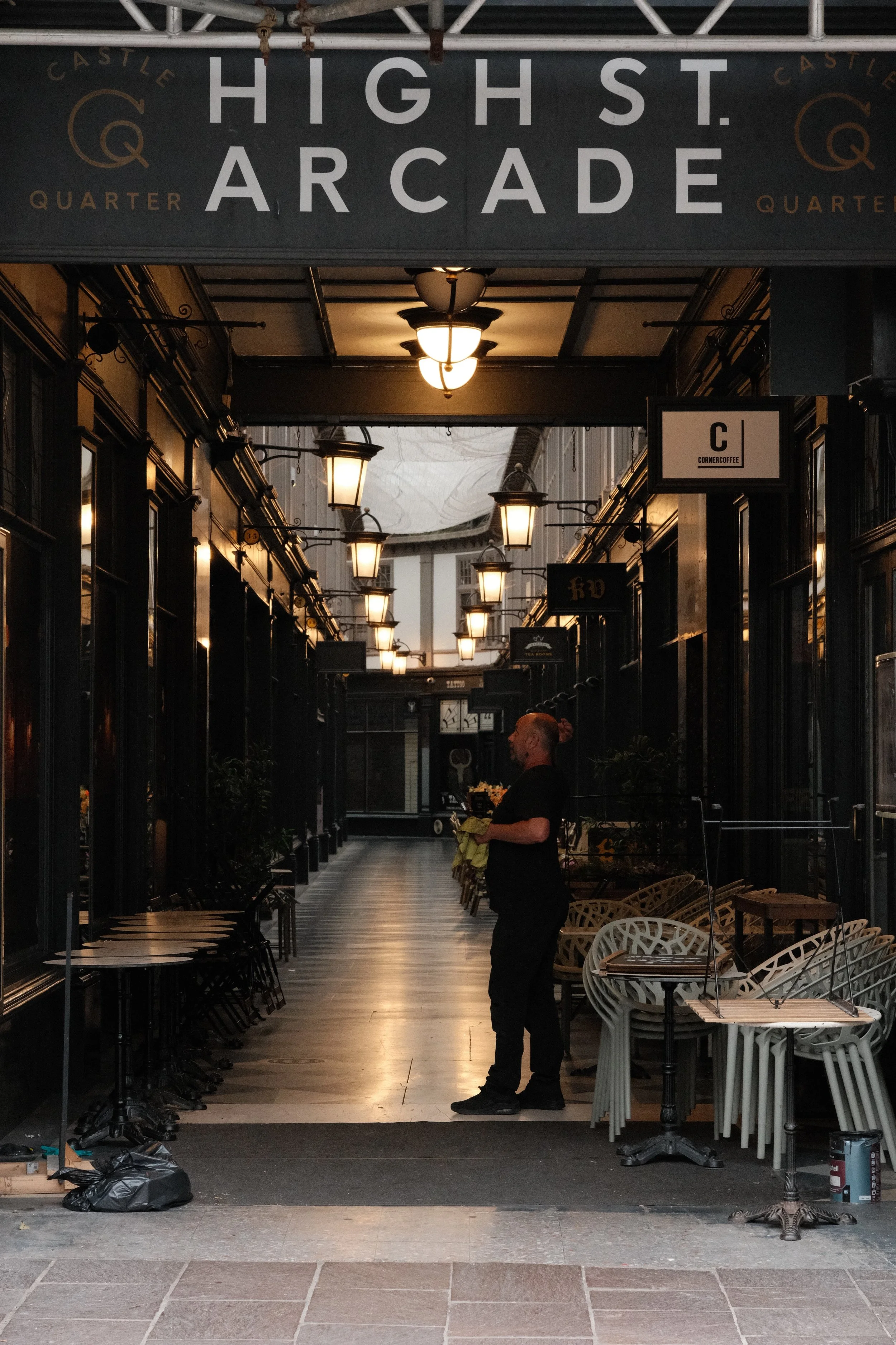 A covered outdoor shopping or dining area with hanging light fixtures, several empty tables and chairs, and a person standing holding a cloth while facing away from the camera. The sign above reads 'High St Arcade'.