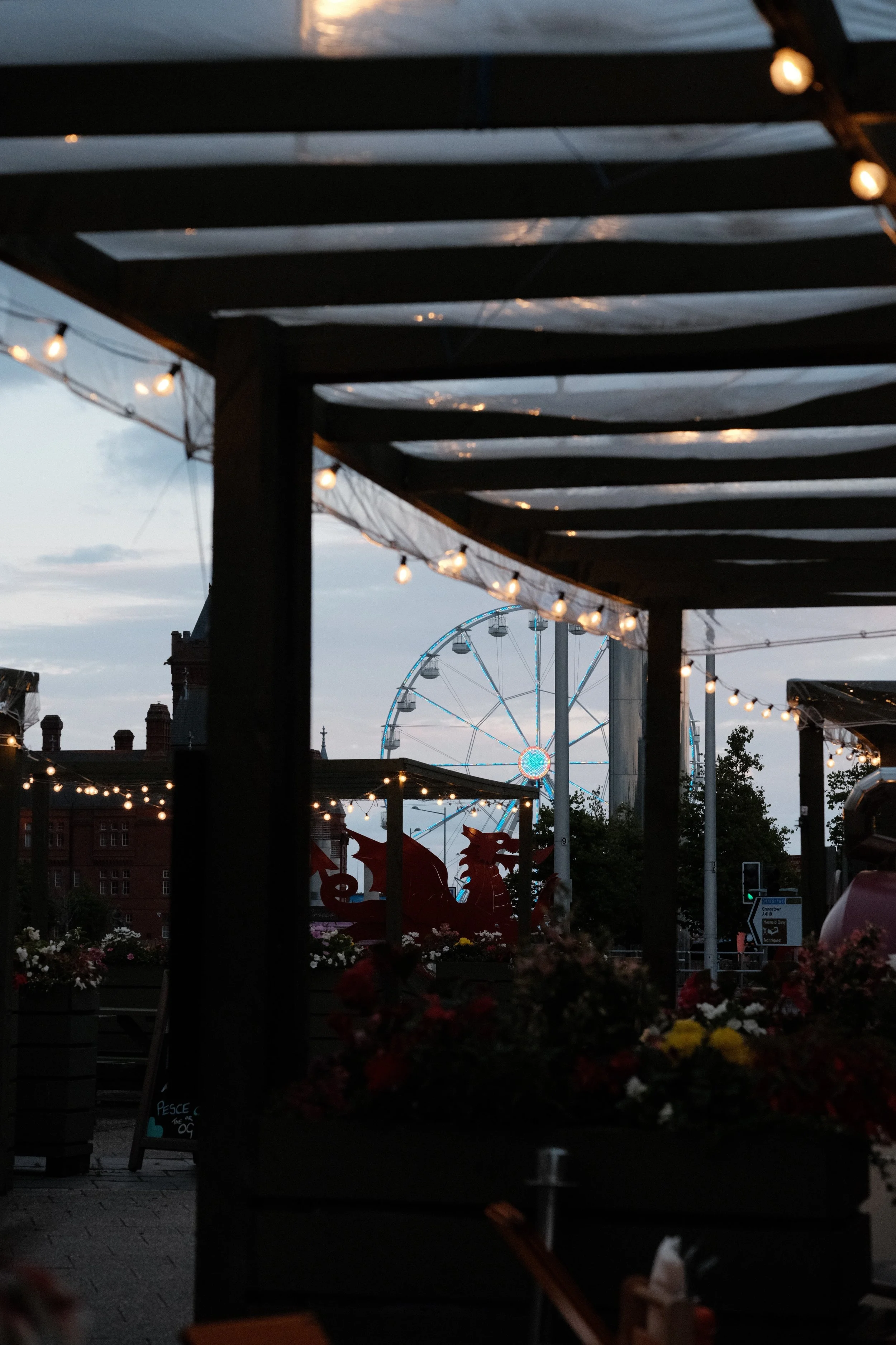 A nighttime scene at a fairground, seen through a wooden structure with string lights. In the background, a large Ferris wheel with illuminated cabins is visible, along with a few dragon-shaped rides and a building with a row of chimneys.