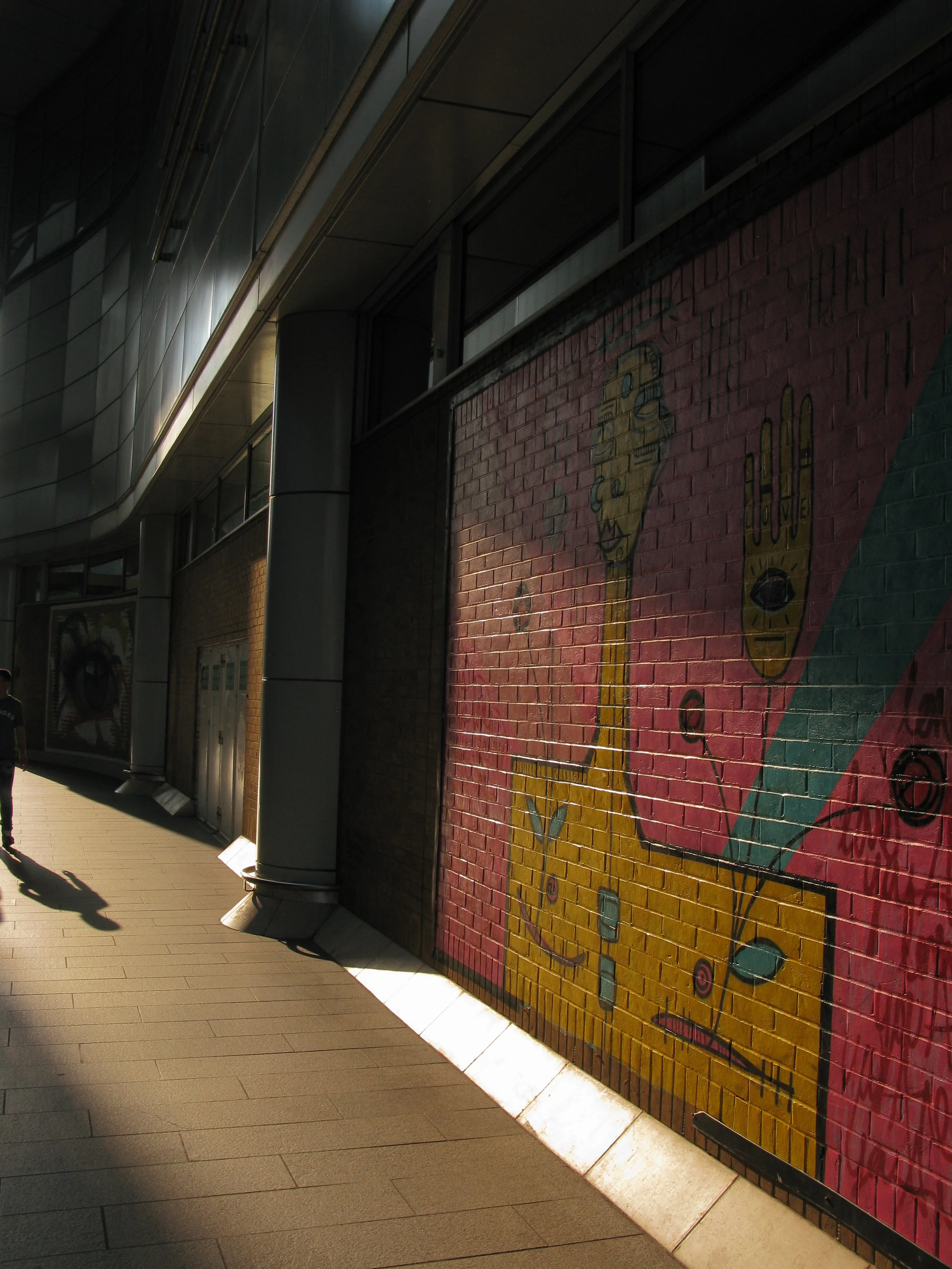 Street view featuring a colorful mural on a brick wall, with a modern building with large windows and a metallic overhang, and a sidewalk with shadows cast by street furniture.