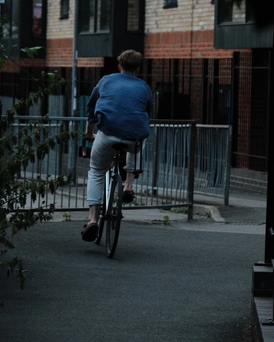 A person rides a bicycle on a city sidewalk during dusk or evening, with buildings and fencing in the background.