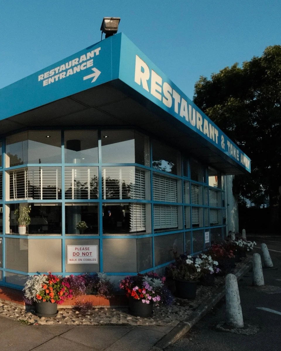 Exterior of a restaurant building with blue signage indicating 'Restaurant & Take Away'. A sign on the window says 'Please Do Not Walk on Cobblestones'. The building has large windows with white blinds, potted flowers outside, and parking stops in th