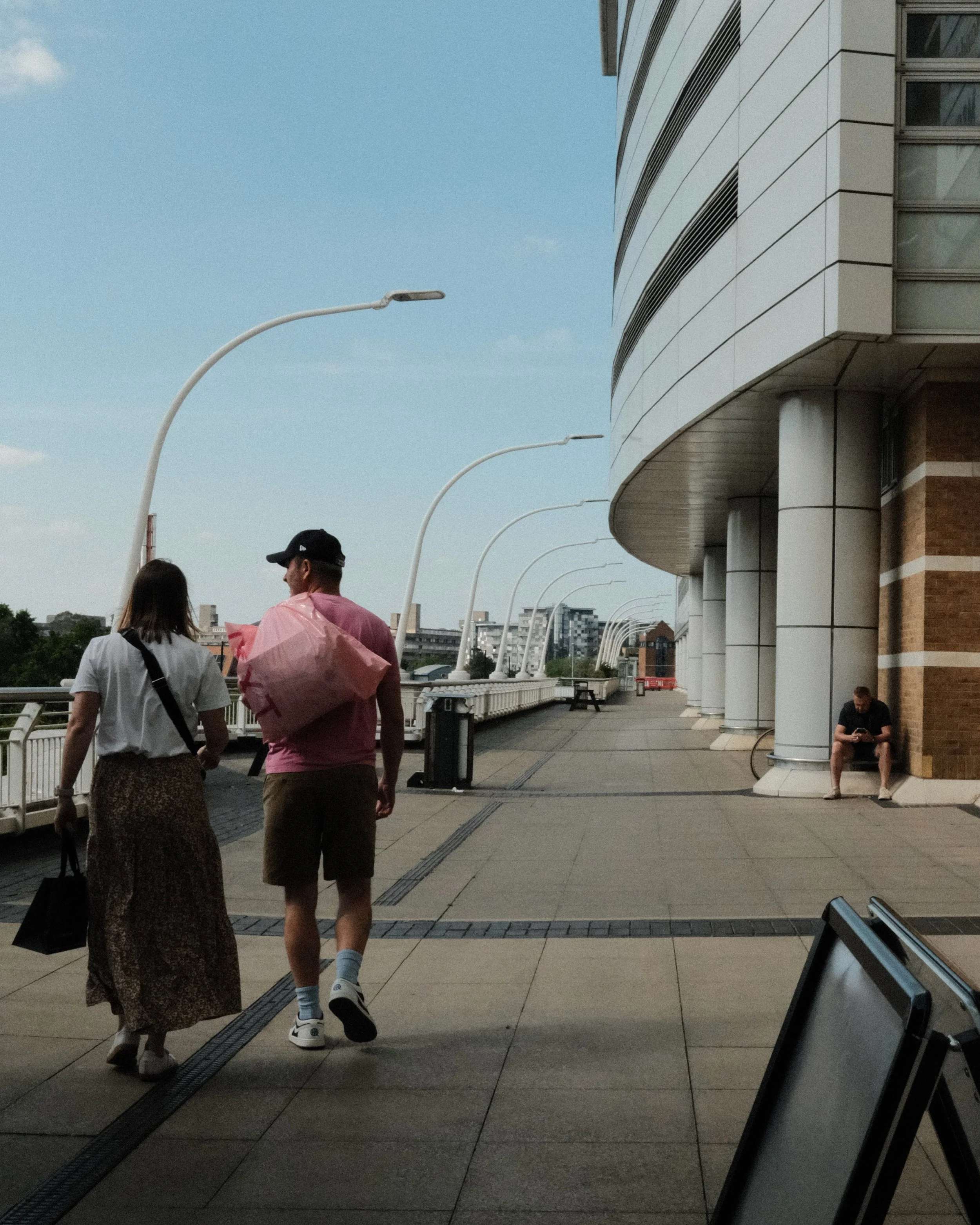 People walking along an outdoor walkway next to a modern building with curved architecture and streetlights, with the sky visible overhead.