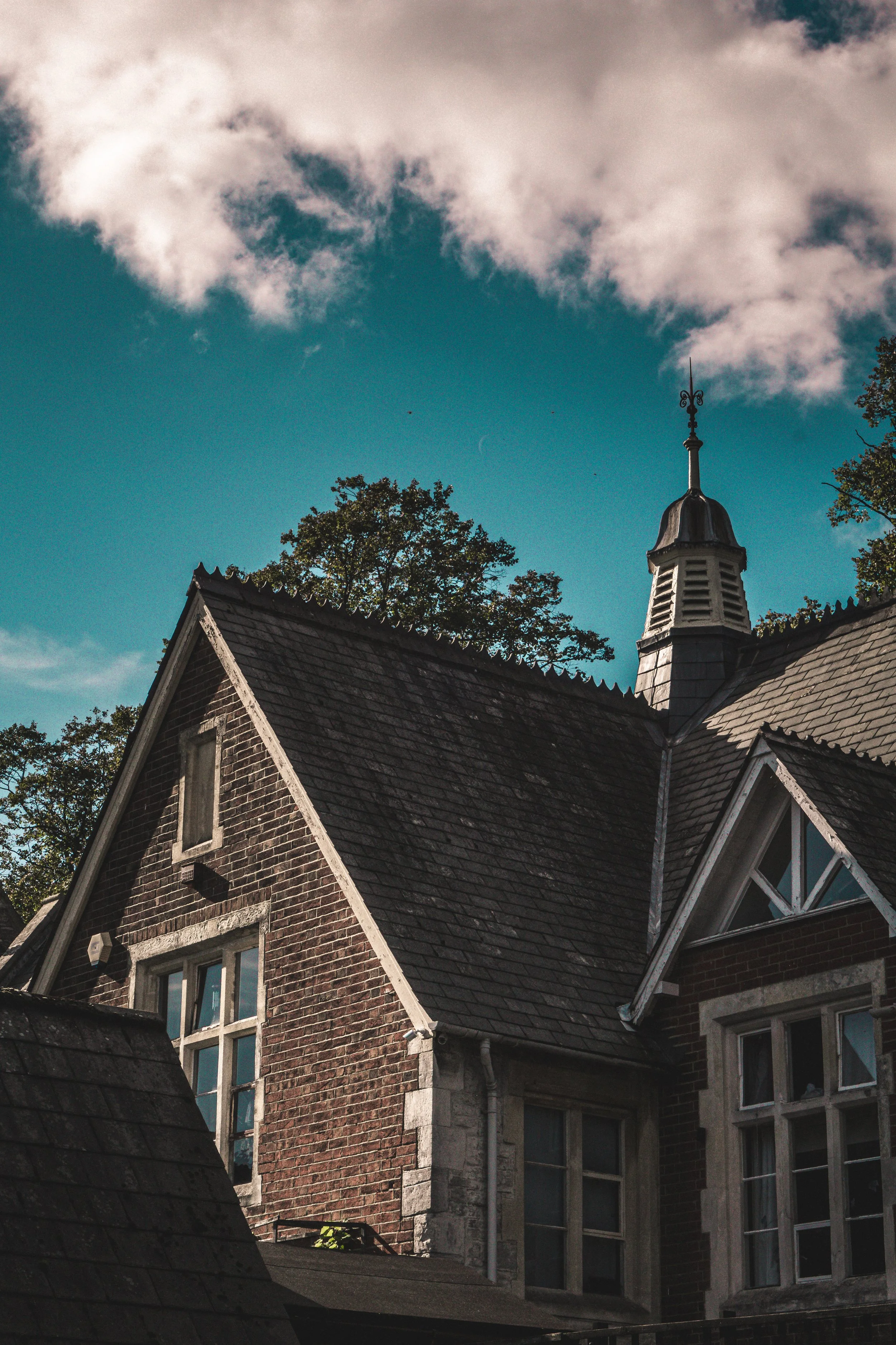 The photo shows the top portion of a historic brick building with multiple windows and a steep roof. There's a prominent ornate spire with metalwork at the highest point of the roof. Trees and a cloudy sky are visible in the background.