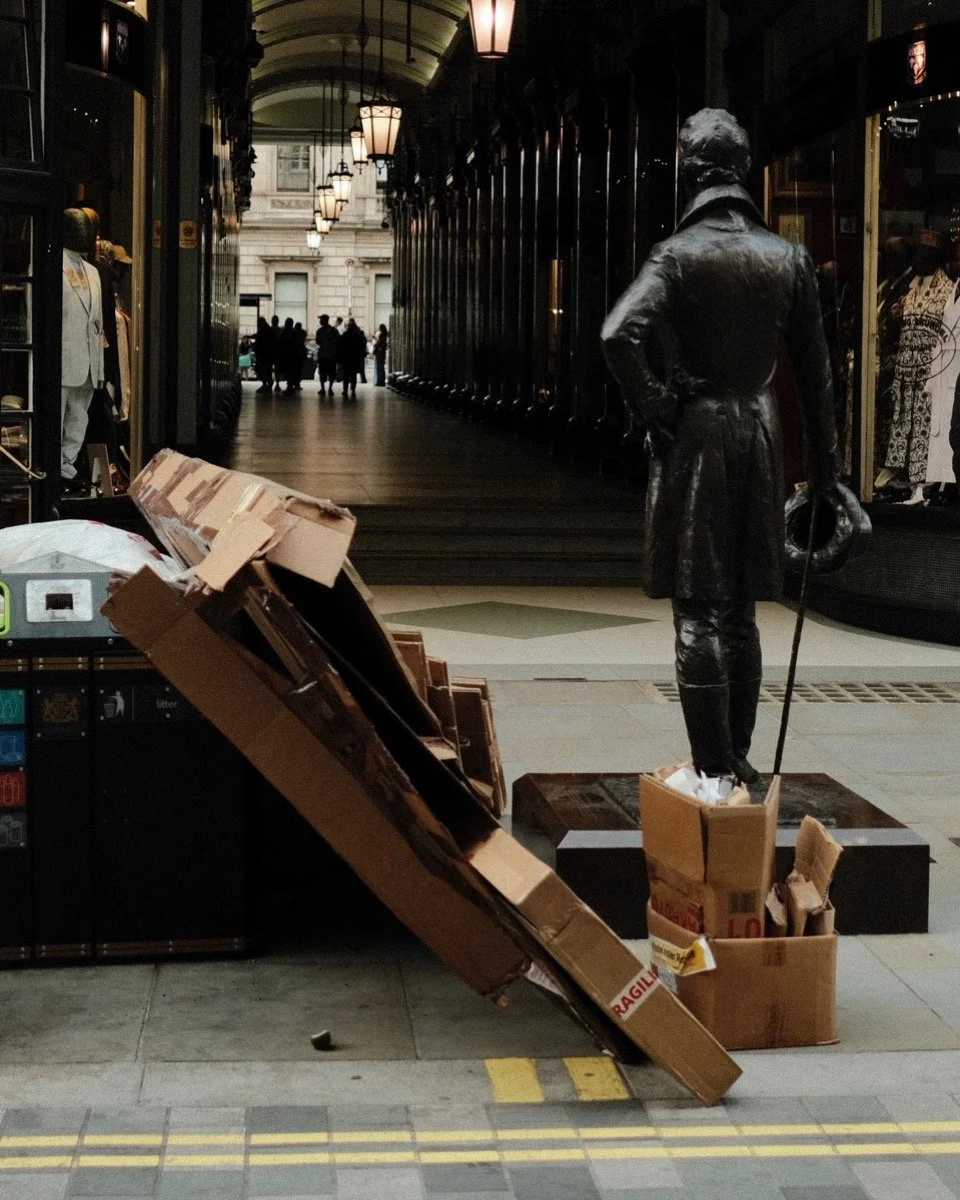 A nighttime city street scene with a statue of a man in a leather coat and hat, standing with his back to the camera, holding a cane and a hat. Nearby are cardboard boxes, one filled with packing material, and a damaged cardboard stand with boxes and