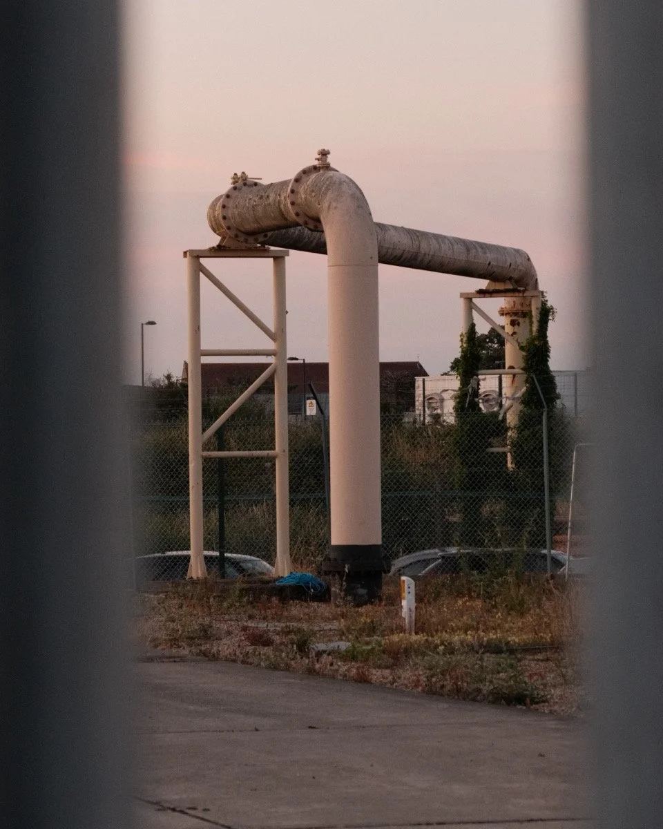 Large industrial pipe structure seen through a fence at dusk, with parked cars and buildings in the background.