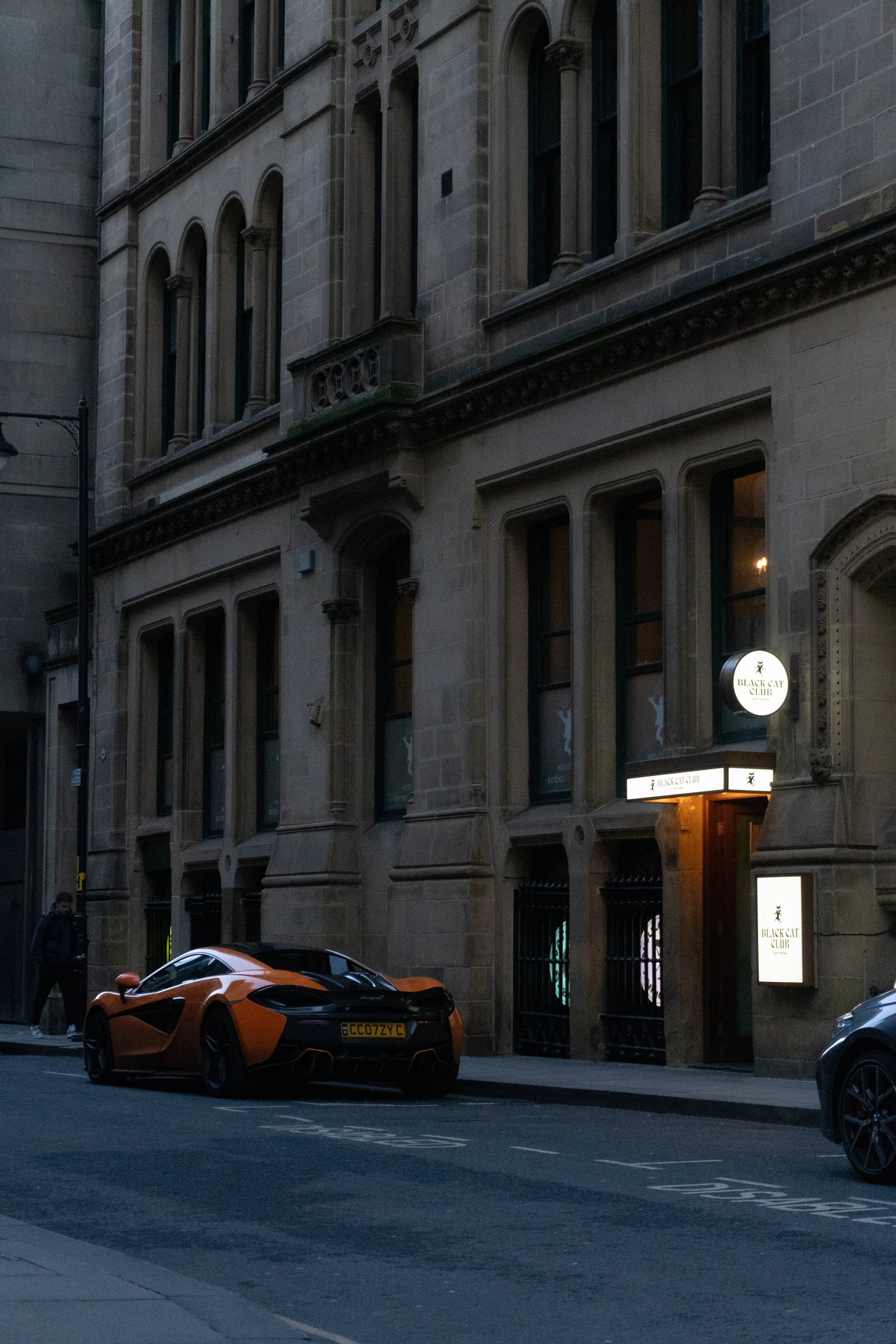 A daytime street scene featuring a luxury orange sports car parked in front of a historic stone building with large windows and ornate architectural details. A person is walking on the sidewalk, and a street lamp is visible on the left side.