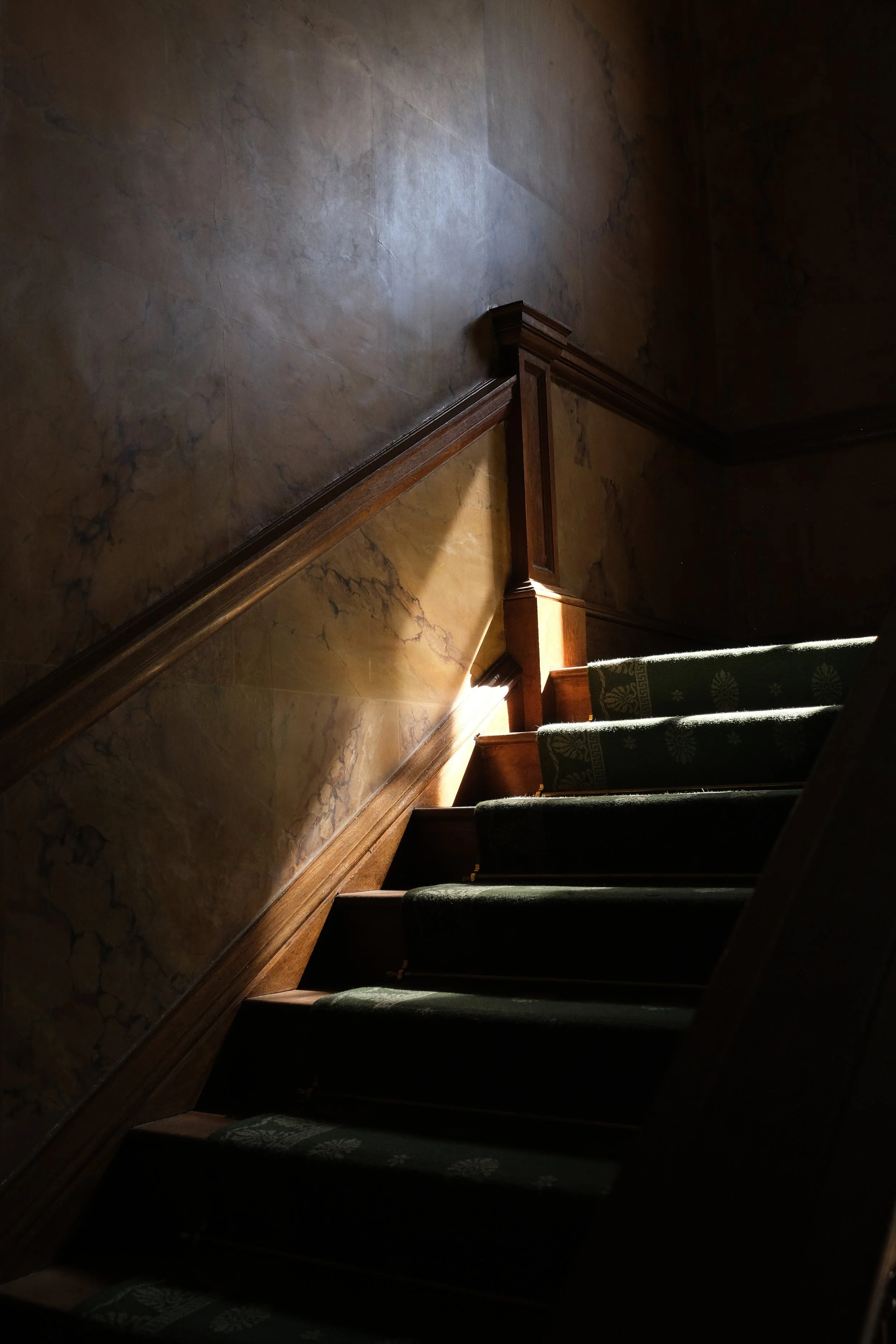Indoor staircase with green patterned carpet runners, wooden trim, and a marble wall with natural light shining through.