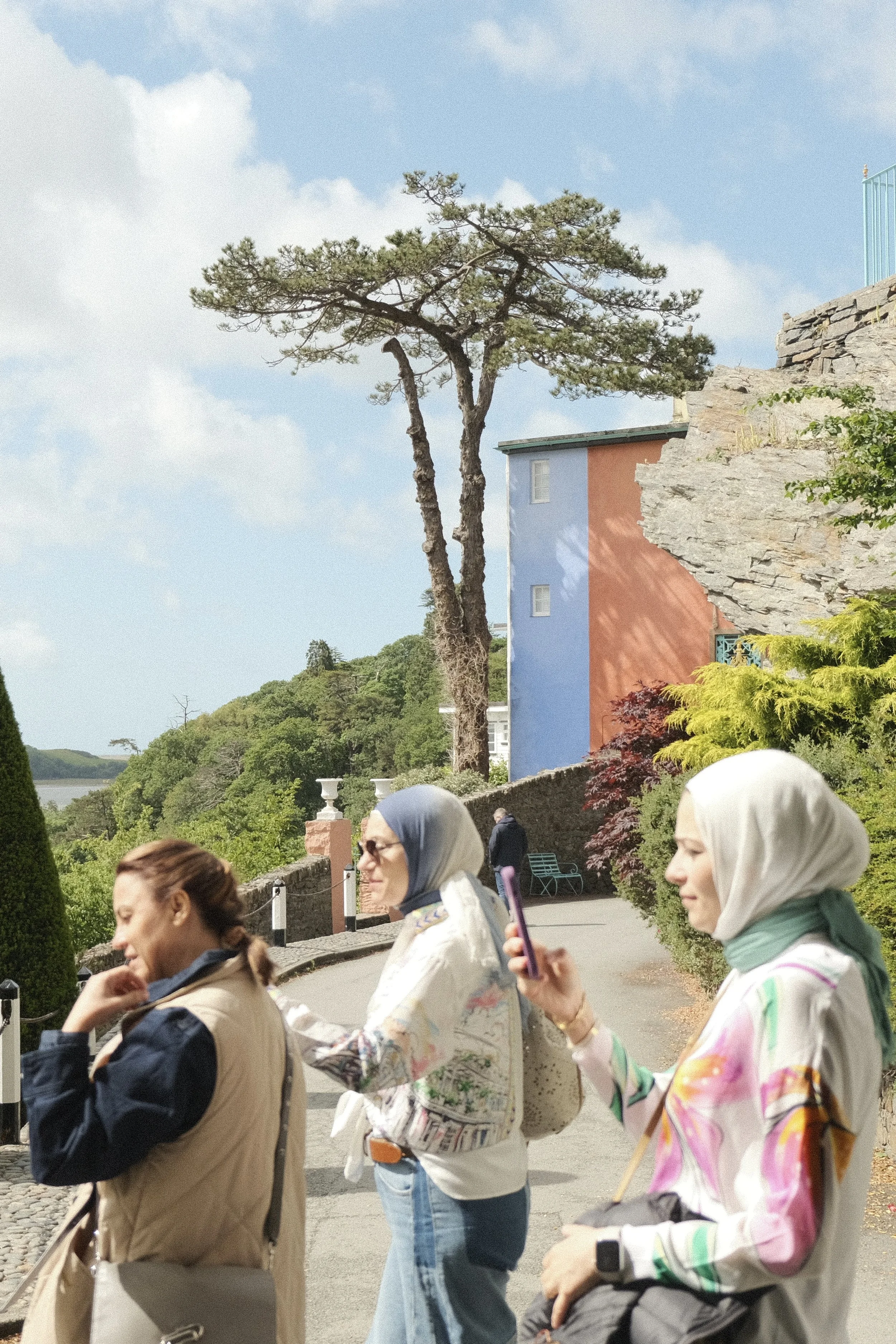 Group of women walking outdoors along a scenic pathway with trees, a small building painted in blue and orange, and rocky formations in the background. Some women are wearing headscarves, and one woman is holding a smartphone.