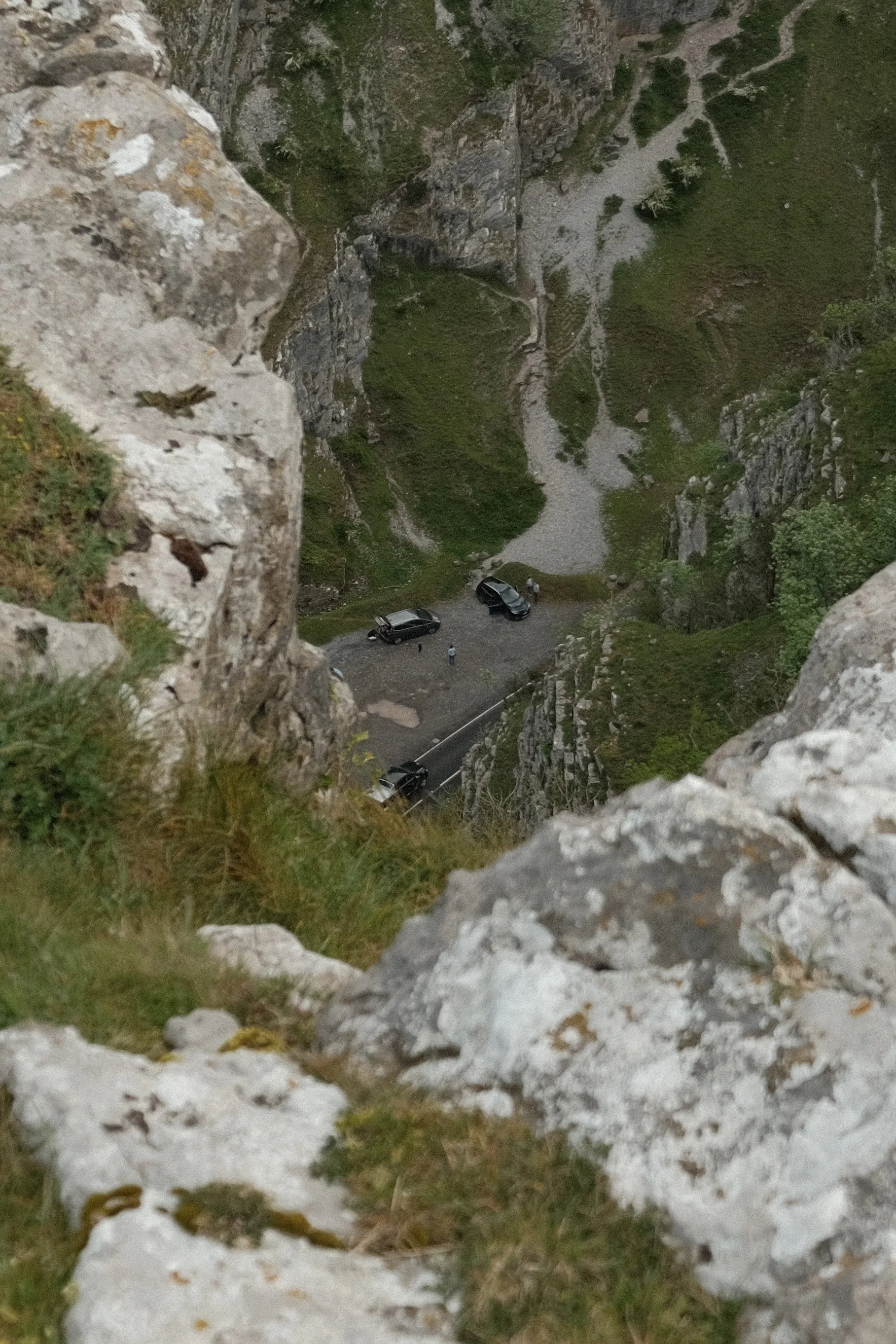 A view from a high point looking down into a rocky, green valley with a small dirt parking area at the bottom where a few cars and people are visible.