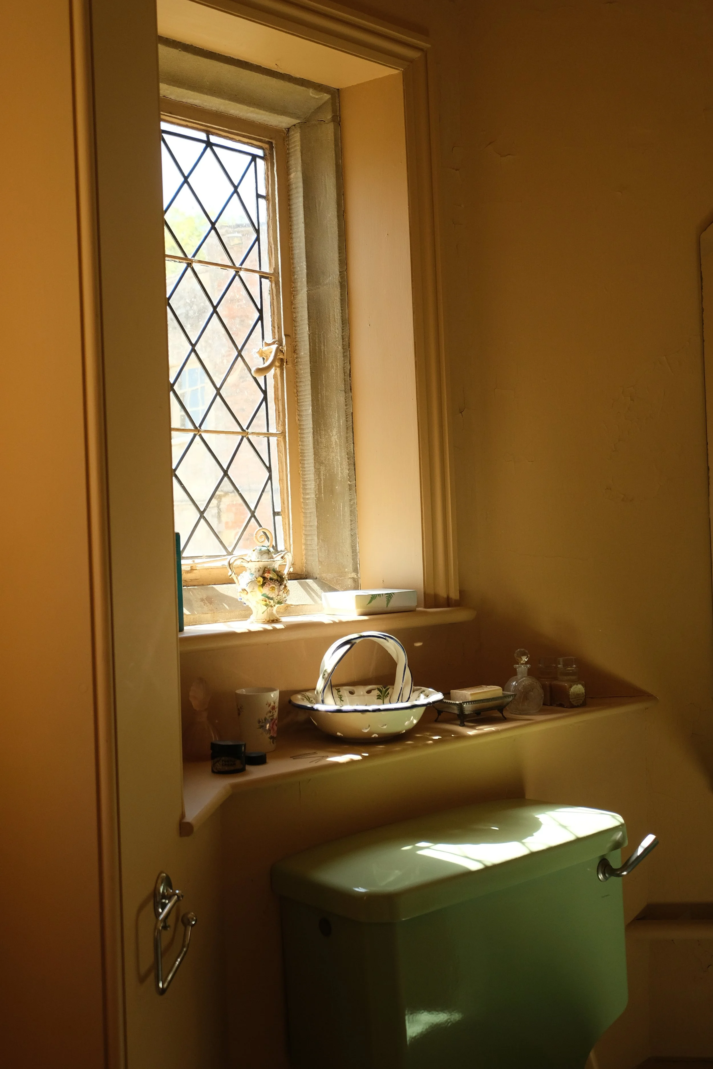 A cozy kitchen window with diamond-patterned glass panes, decorated with a floral teapot, a small box, and jars beneath it, sunlight illuminating the scene, with a vintage green trash bin below the window.