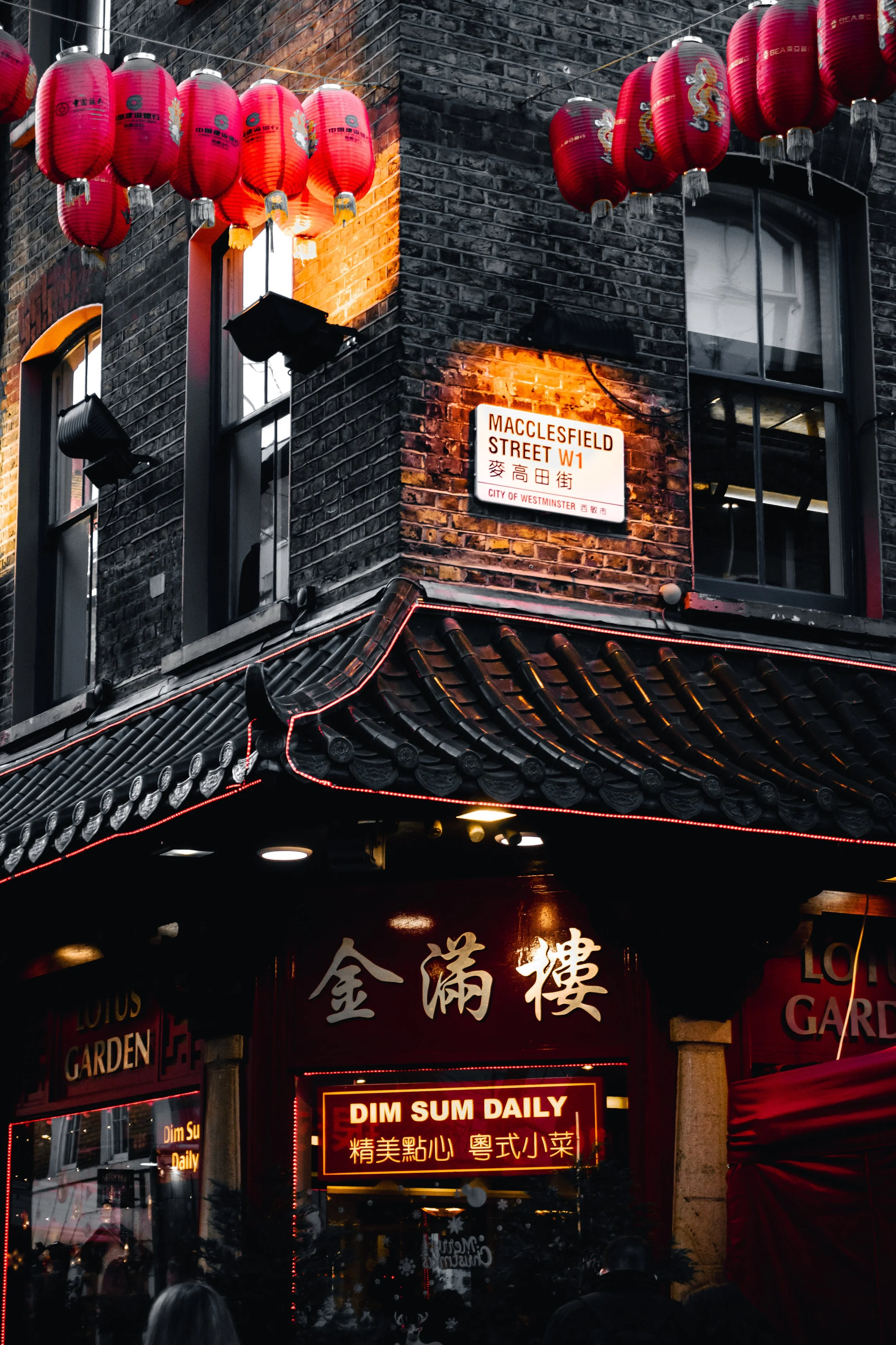 Red Chinese lanterns hanging above a street in Chinatown, with a neon sign advertising dim sum daily and a street sign for Macclesfield Street in Westminster, London. The scene is bustling with people and illuminated by decorative lights.