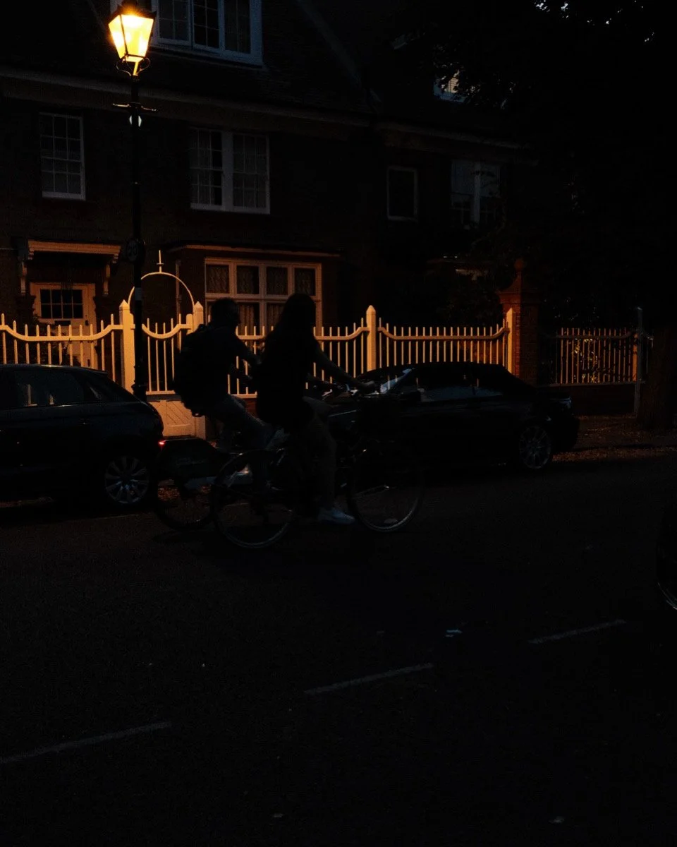 Two people riding a bicycle at night near parked cars and a streetlamp on a dark residential street.
