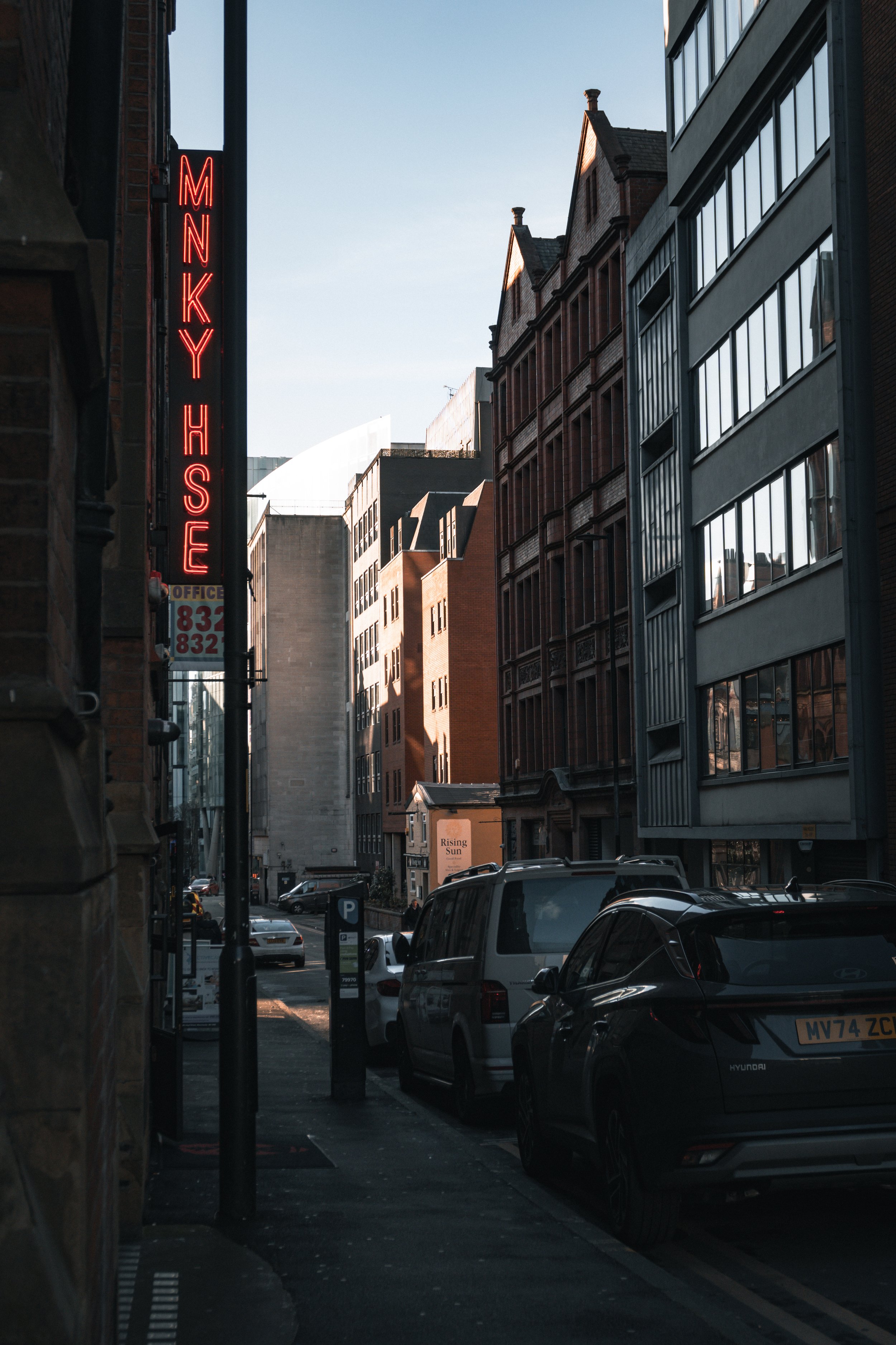 A city street scene with parked cars on the right side, and tall buildings on either side. A red neon sign on the left spells out 'MNKY HSE'. The sky is clear with some sunlight illuminating the tops of the buildings.
