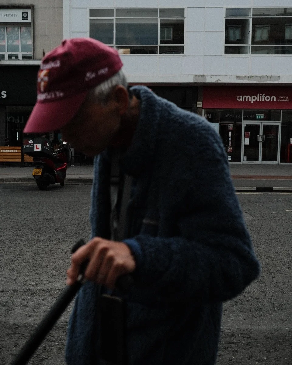 A man wearing a maroon cap and a blue sweater, holding a cane or walking stick, standing on a city street with shops and a scooter in the background.