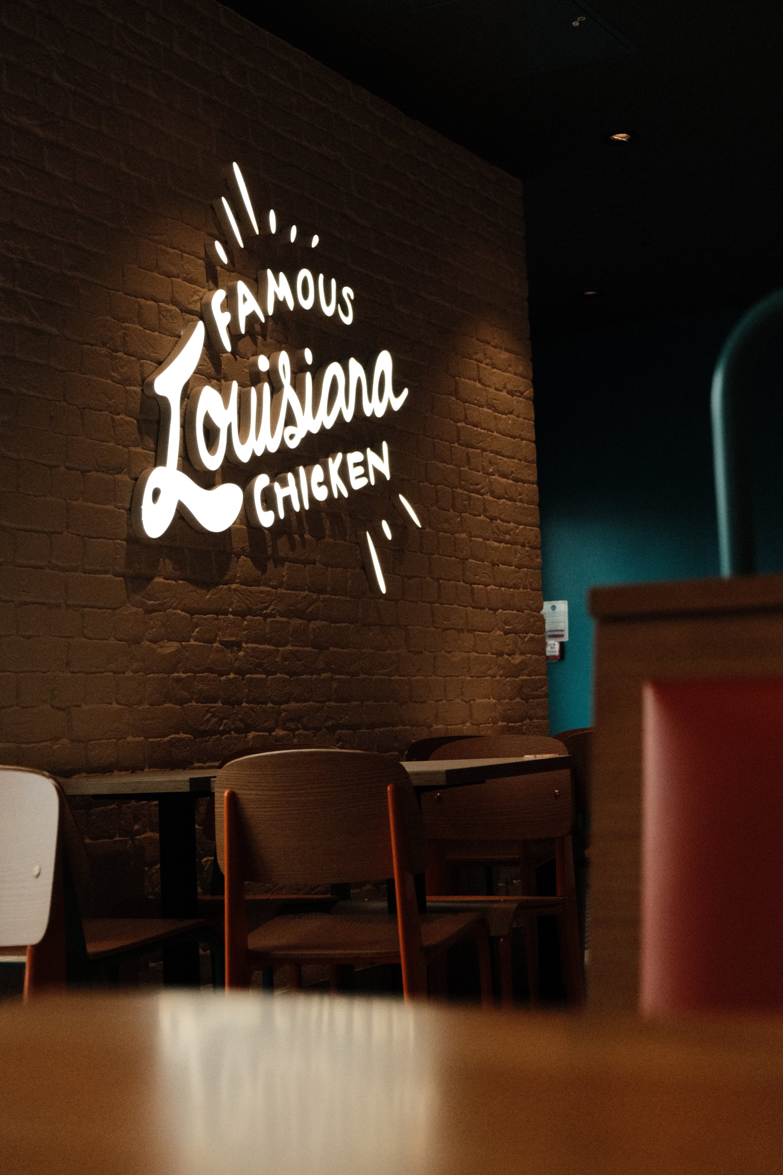 Neon sign reading 'Famous Louisiana Chicken' on a brick wall inside a restaurant, with chairs and tables in the foreground.