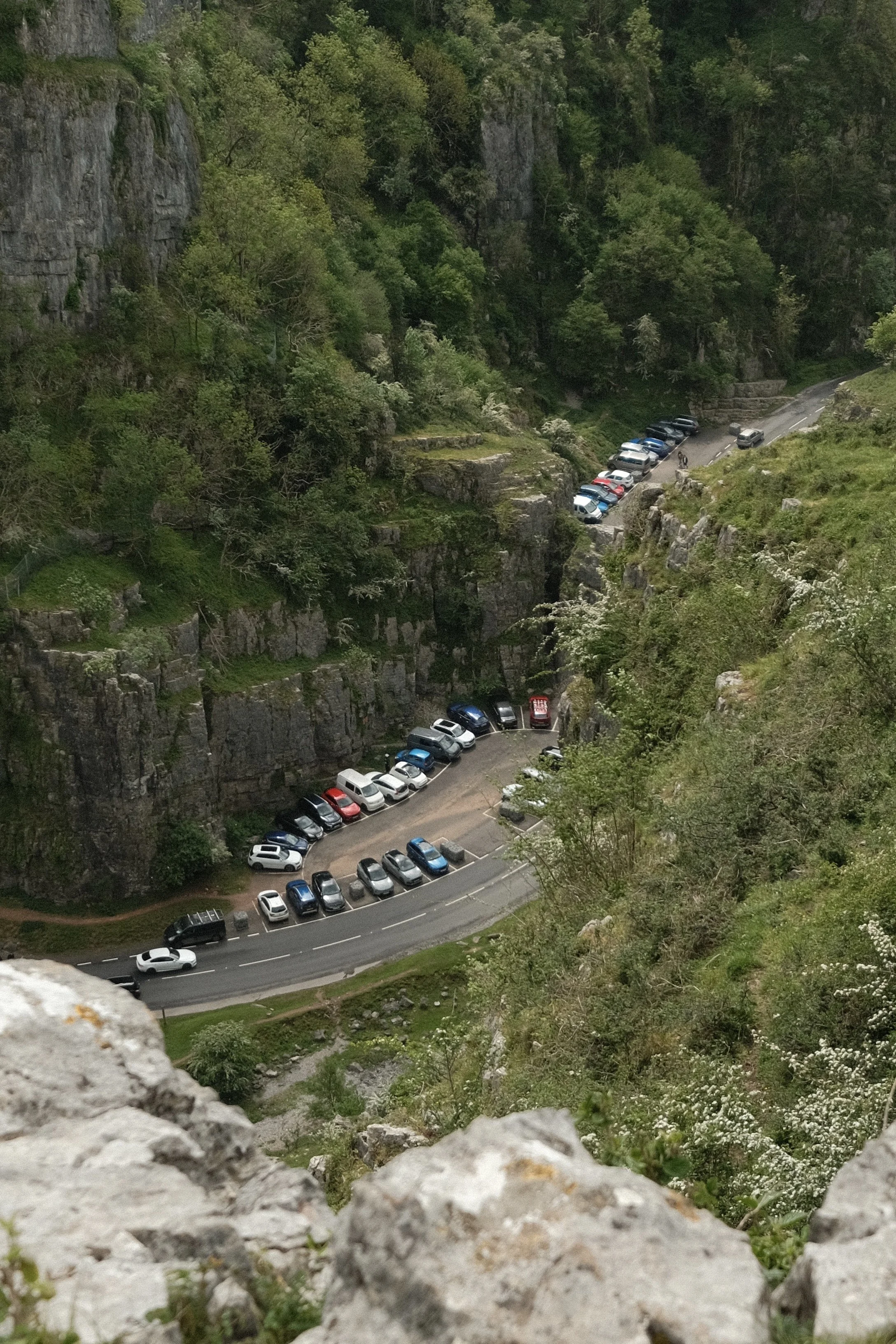 A winding mountain road with multiple vehicles parked alongside the edge of a steep, rocky hillside covered in green trees and bushes.