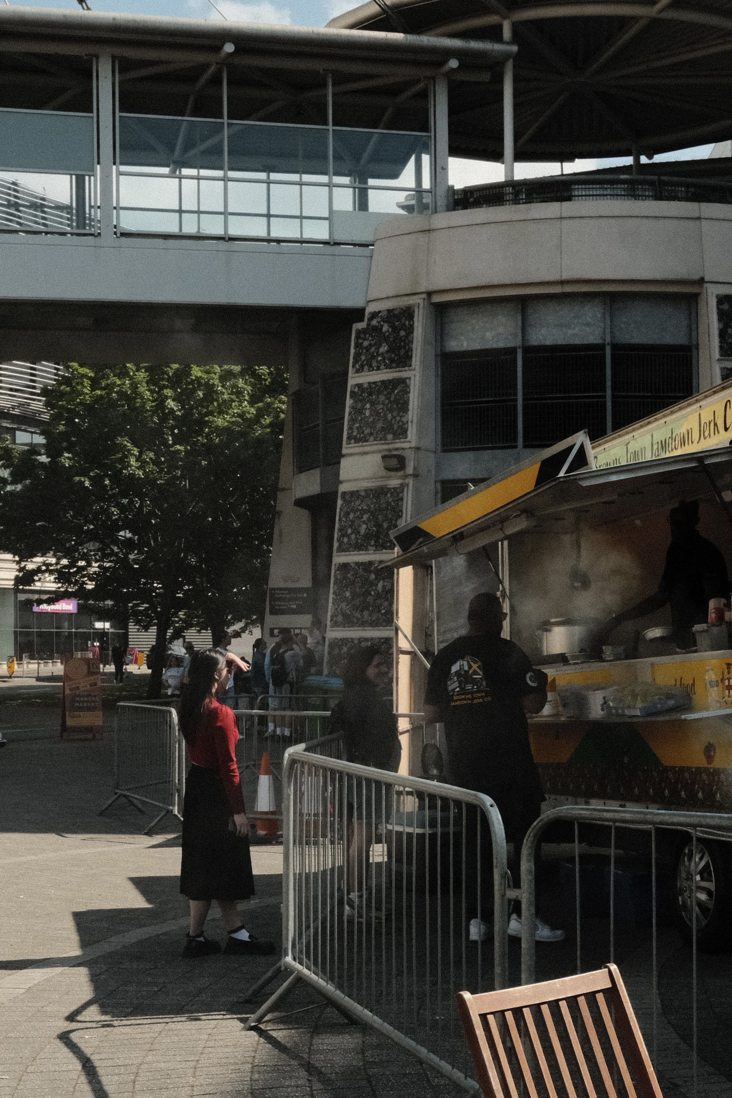 A food truck serving food at an outdoor event with a woman standing nearby and a few people in line in front of the truck. There are barriers, trees, and a building in the background.