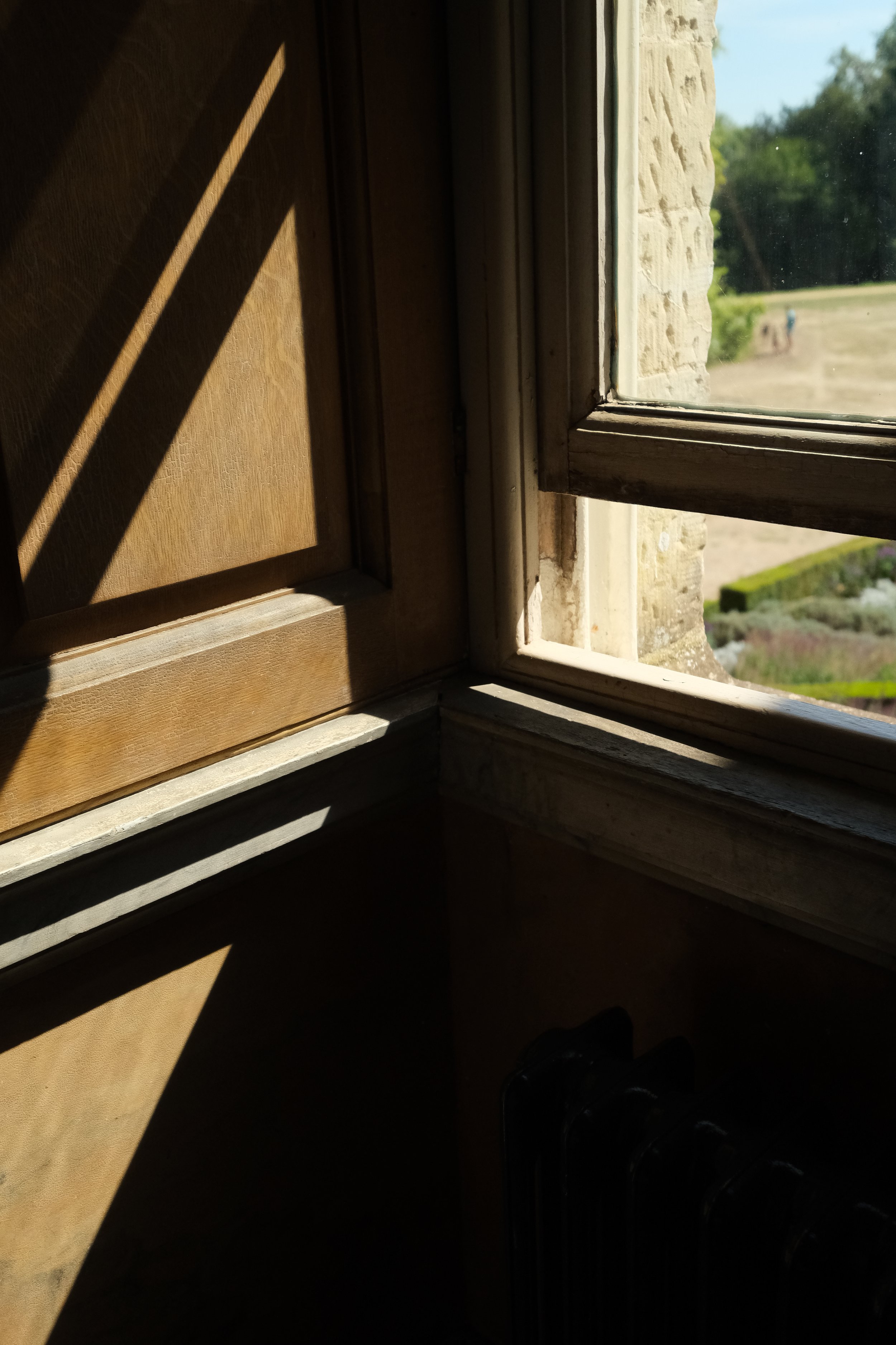 View through a window showing a garden and people walking outdoors, with sunlight casting shadows and a brick wall on the side.