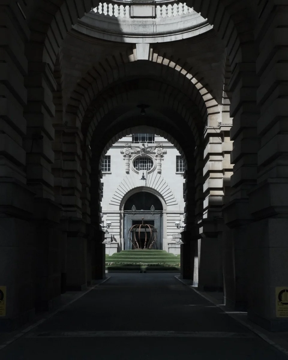 Stone archway leading to a building with a metal sculpture in front, steps, and grass.
