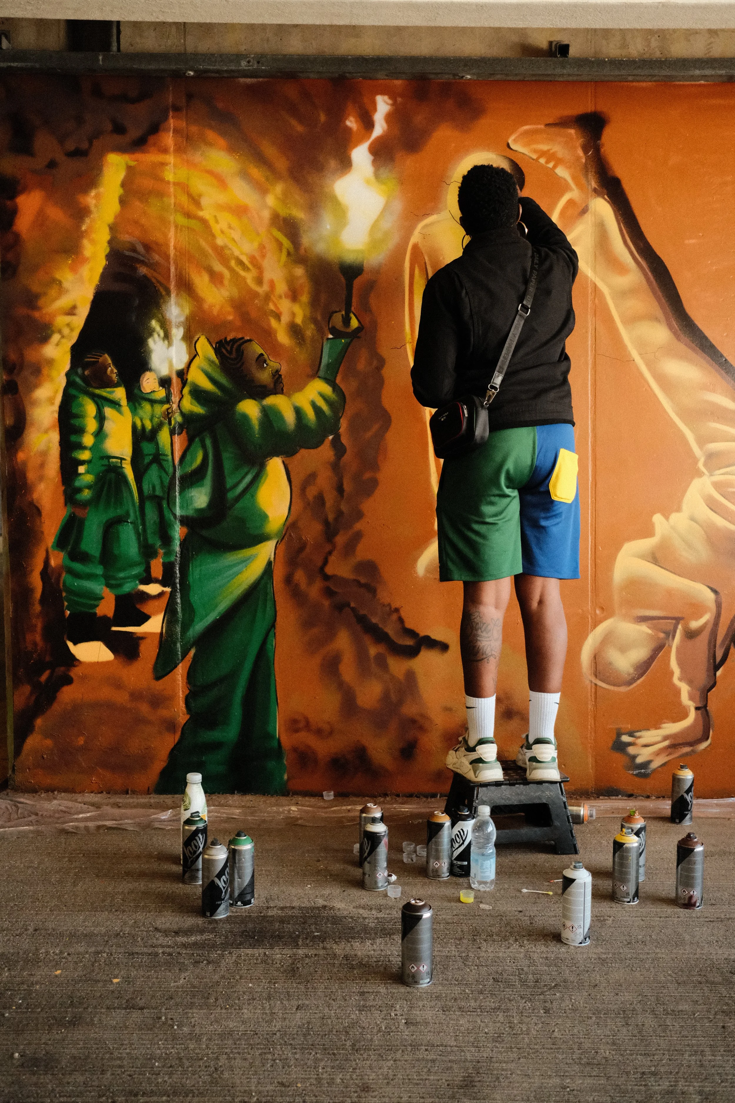 A street artist spray painting a mural featuring three children holding torches, with one child in the foreground holding a lit torch. The artist is wearing shorts, a black jacket, and white sneakers, standing on a small stool. Spray paint cans are s