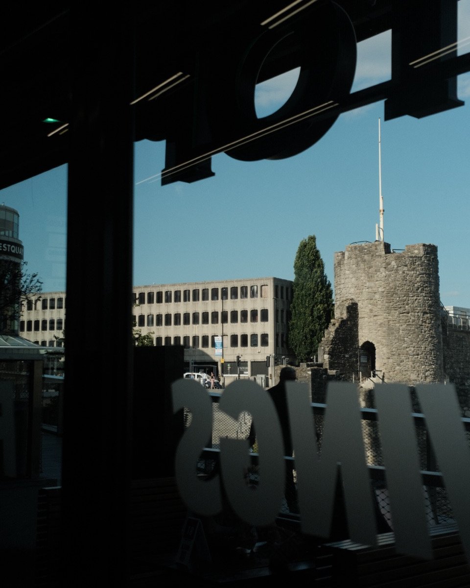 View of a castle tower and modern buildings through a window, with reflections of indoor lights and a sign partially visible.