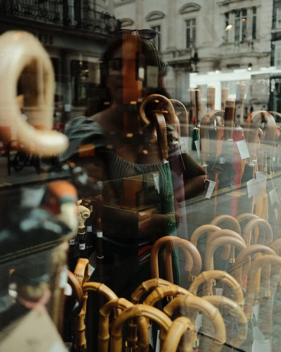 Reflections of a woman wearing sunglasses and a green top, looking through a window display of umbrellas and walking sticks outside a building.